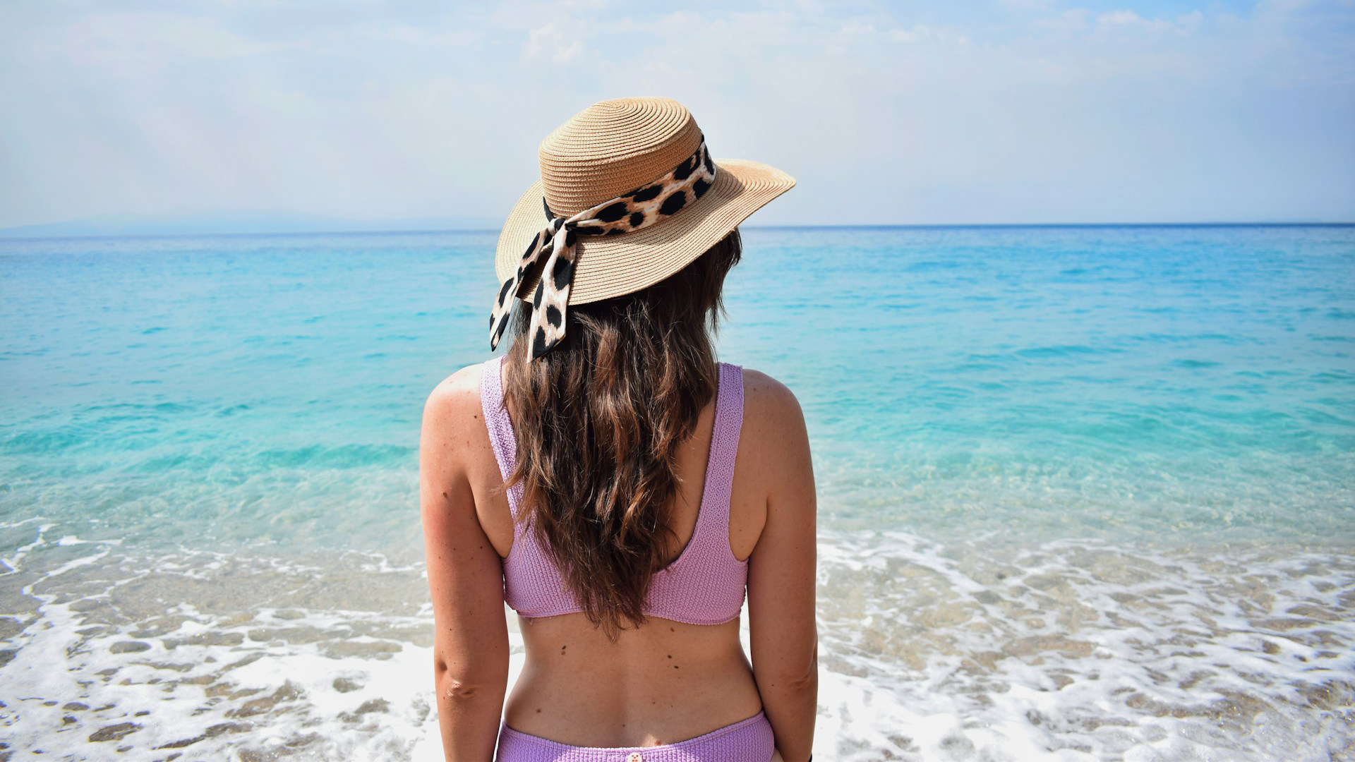 Vrouw in een paarse bikini en hoed met luipaardprint lint, kijkt uit over een heldere blauwe zee vanaf het strand.