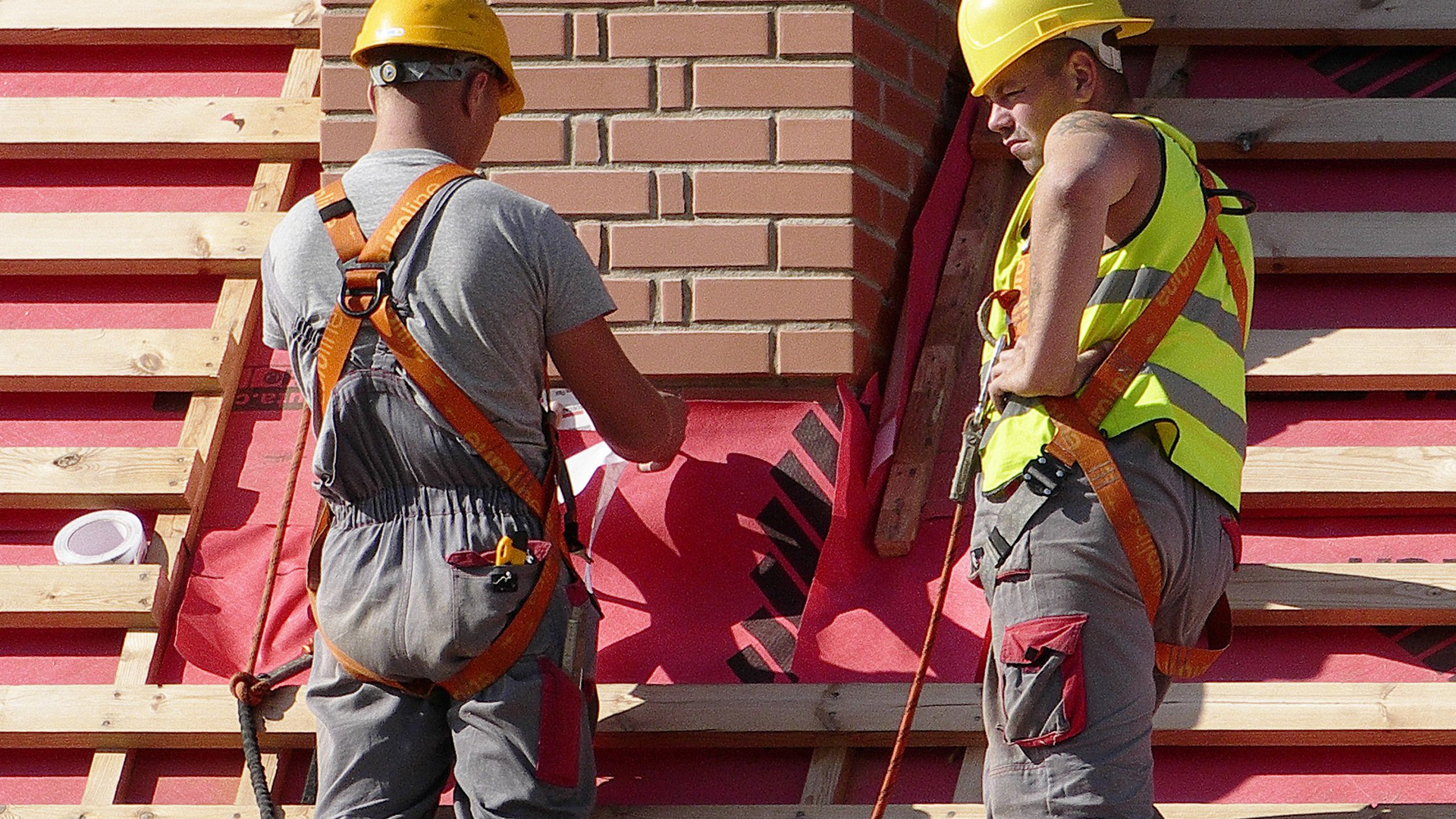 Twee arbeiders dragen veiligheidsuitrusting en helmen terwijl ze buiten aan bouwconstructie werken.