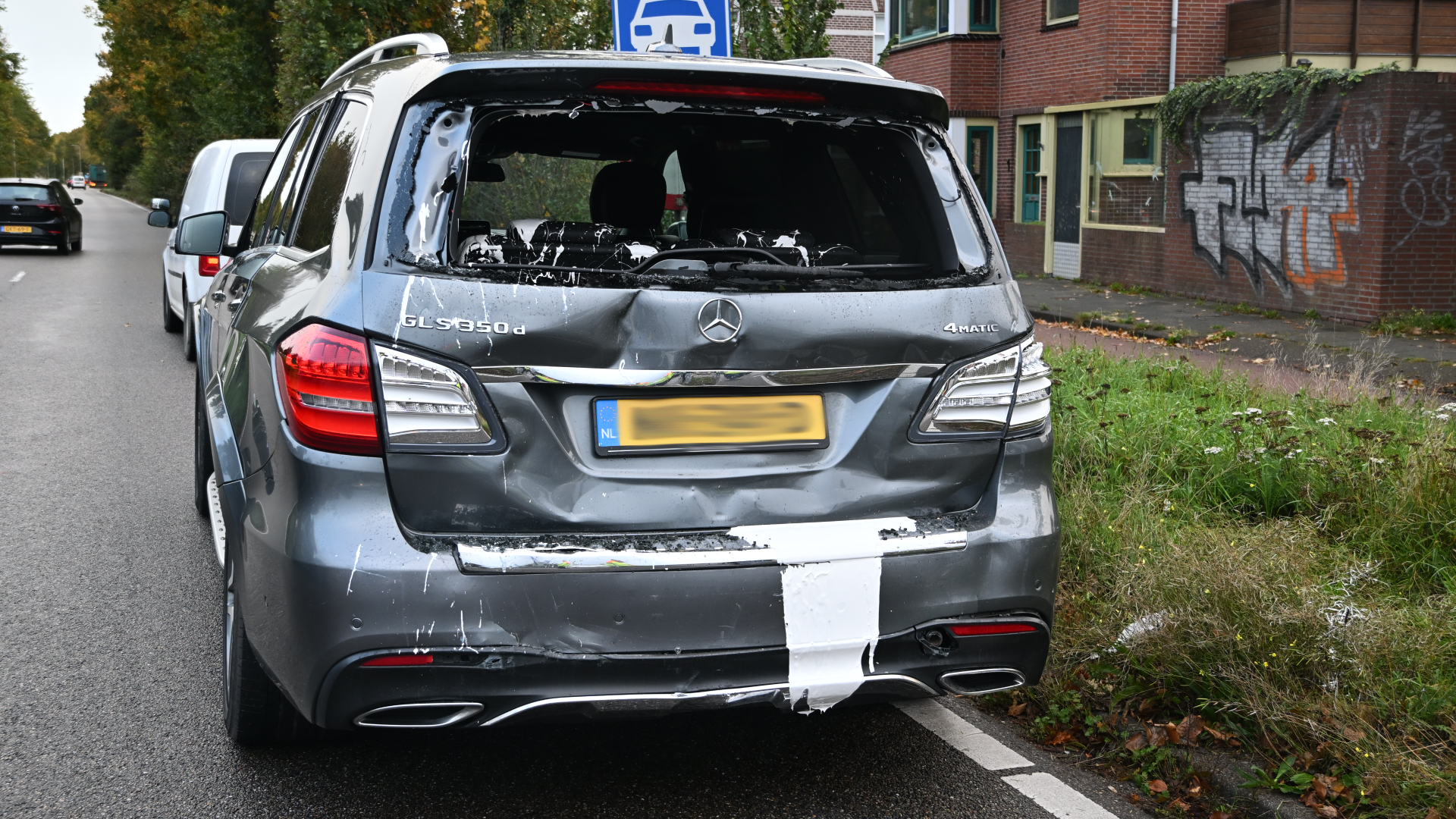 A Mercedes SUV with a damaged and painted rear, parked on a street next to a grassy area and a brick building with graffiti.