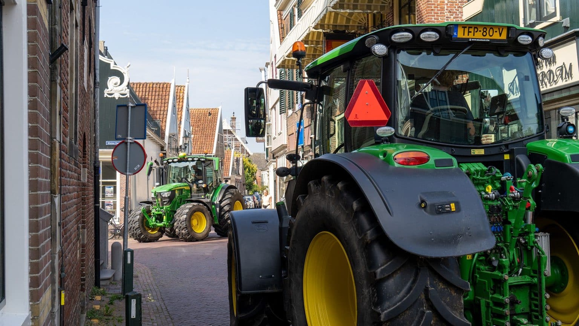 Twee groene tractoren rijden door een smalle stadsstraat met bakstenen gebouwen aan weerszijden.