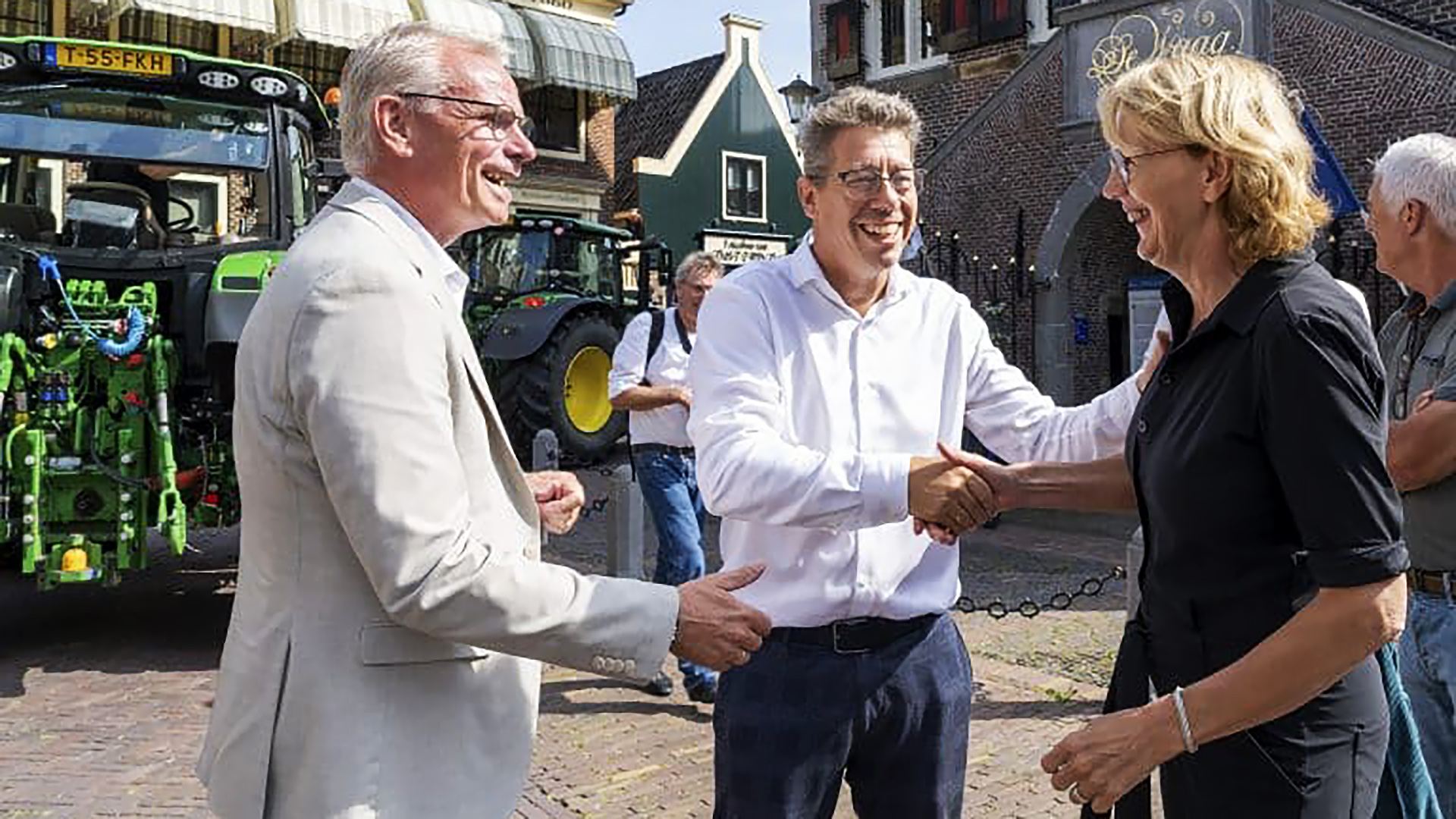 Drie mensen, twee mannen in lichte kleding en een vrouw in een zwarte blouse, schudden elkaar lachend de hand op een plein met een tractor op de achtergrond.