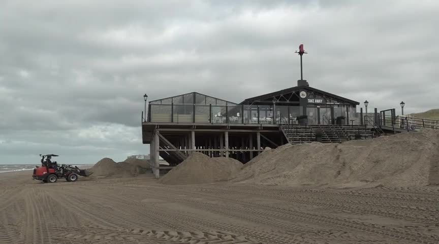 Een strandpaviljoen op verhoogde palen met grote glaspartijen, gelegen naast zandduinen, met een graafmachine geparkeerd op het strand ernaast.