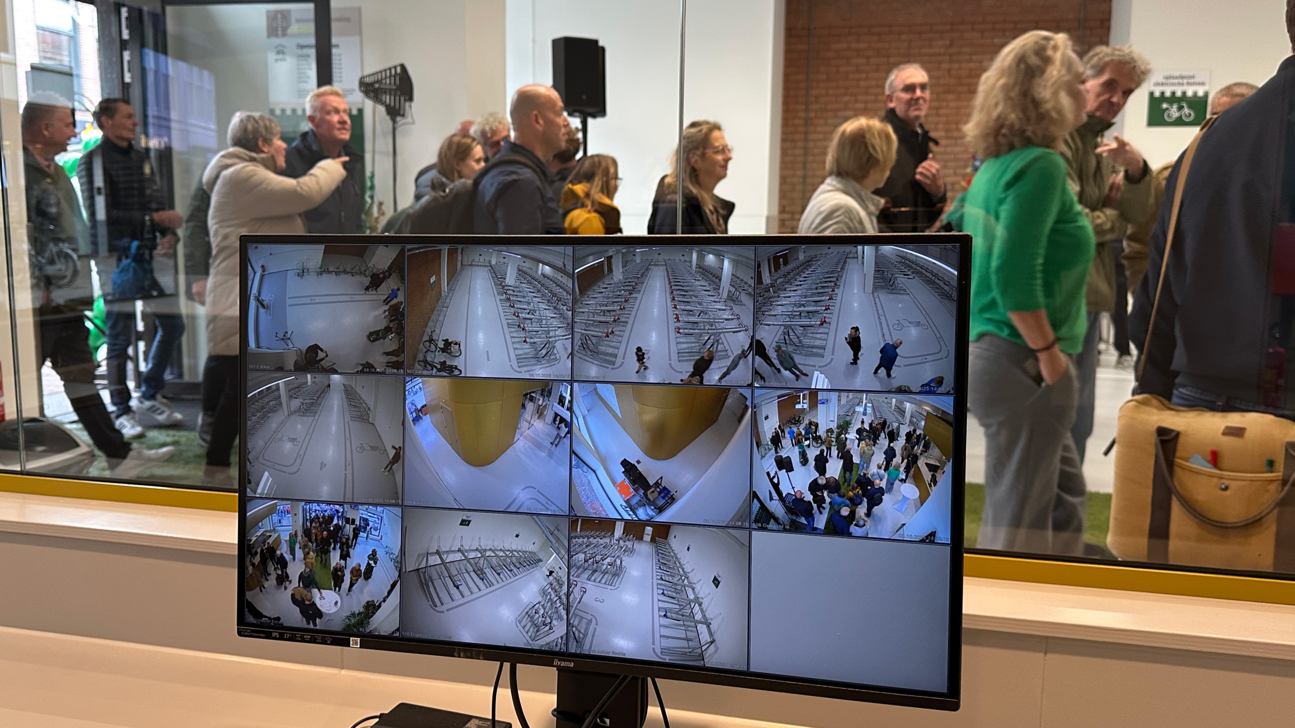 Een groep mensen staat voor een glazen wand, waarachter een scherm met camerabeelden van een fietsenstalling te zien is.