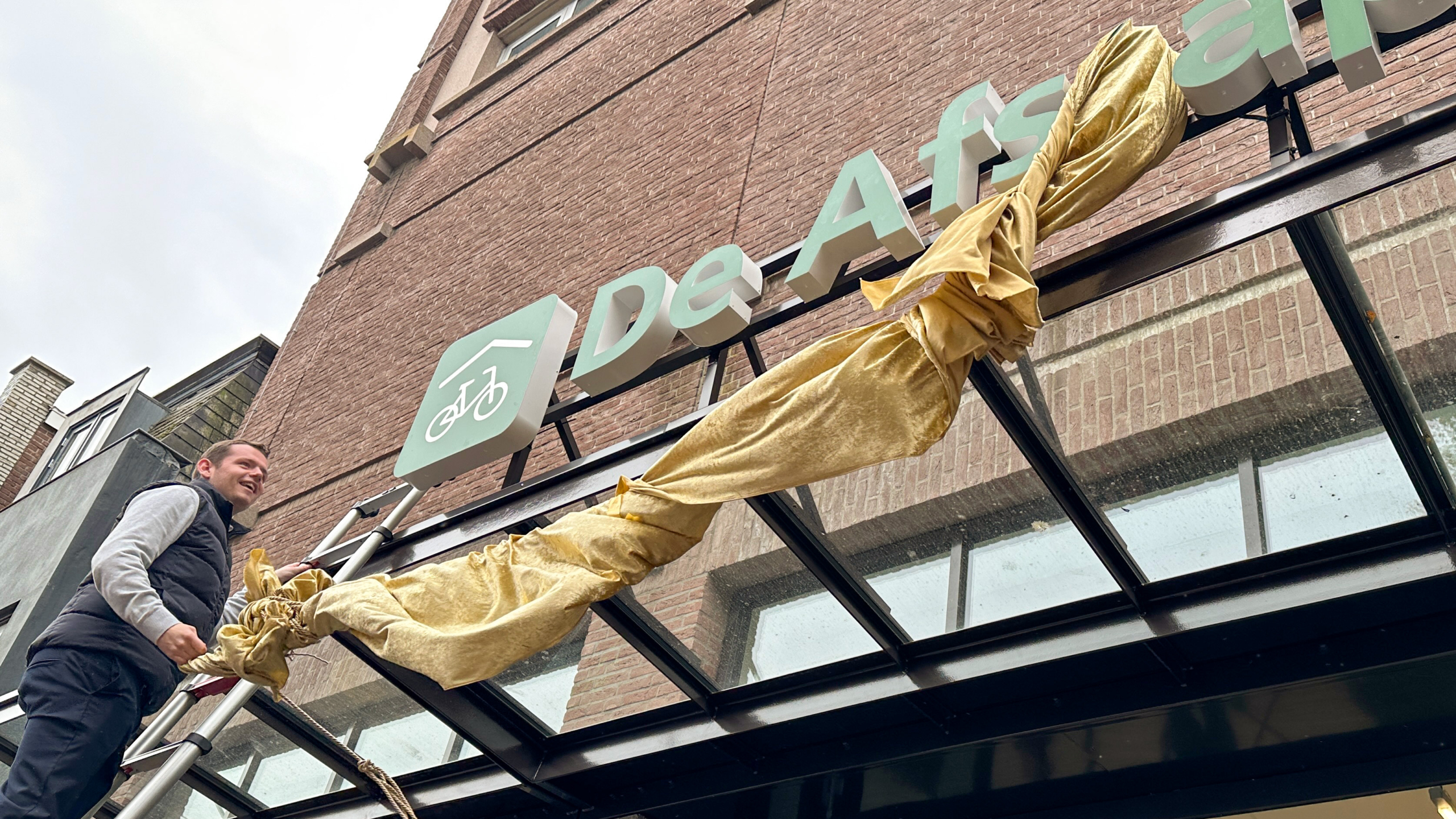 Een man op een ladder onthult samen met een goudkleurig doek een gevelbord met een fietslogo en de tekst 'De Afstap'.