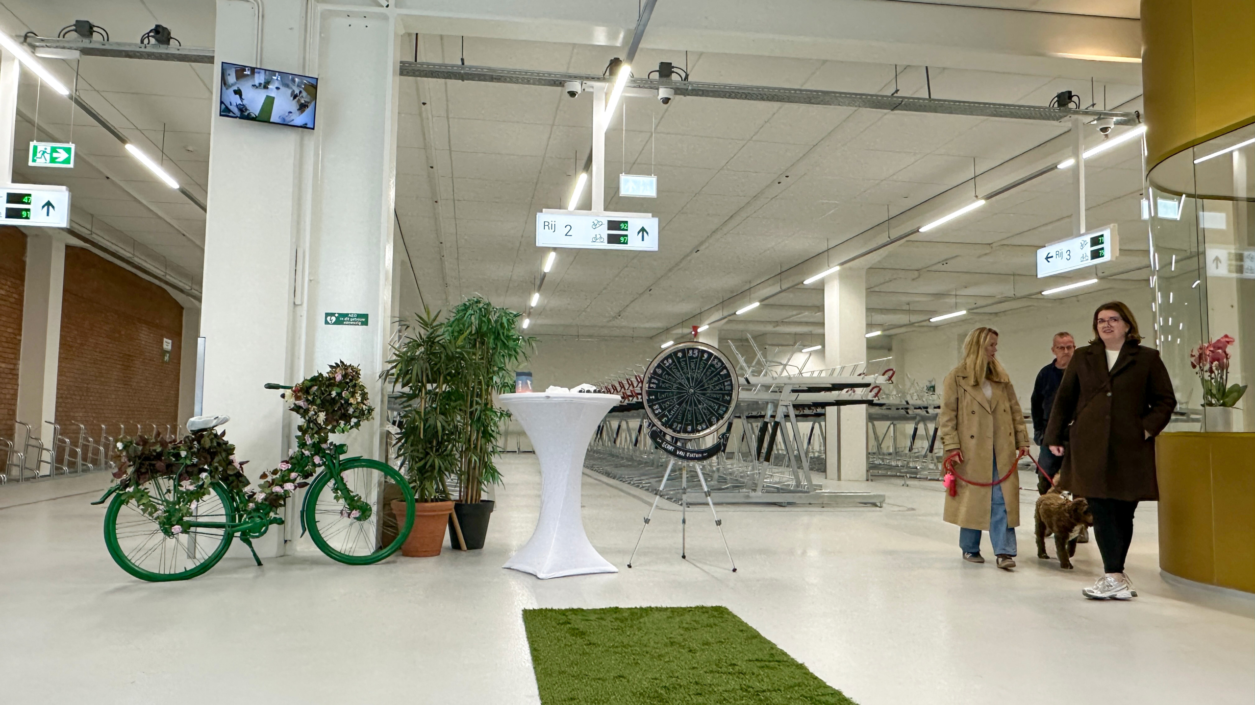 Binnenruimte met een groene versierde fiets, een tafel met een wit kleed en een draaischijf, met mensen en een hond die langs een fietsenstalling lopen.