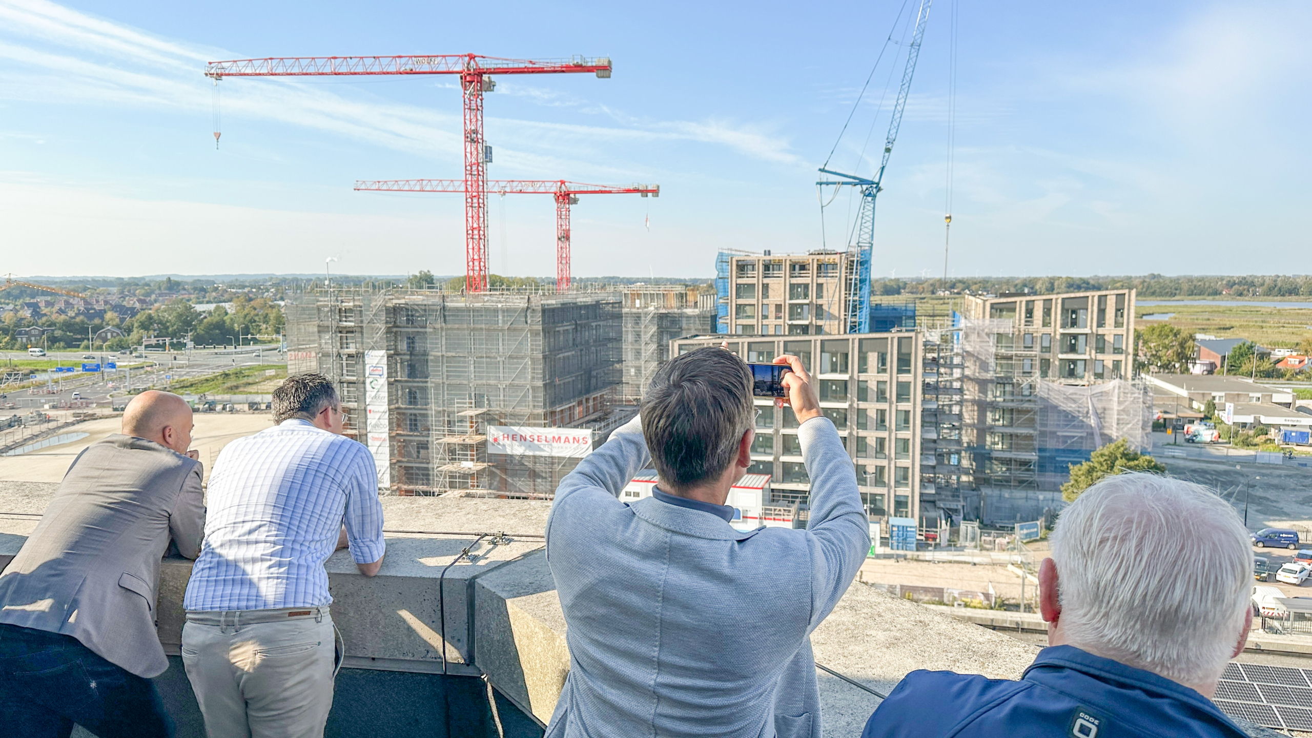 Mannen kijken vanaf een hoogte naar een bouwplaats met hijskranen en gebouwen in aanbouw, met één persoon die een foto maakt.
