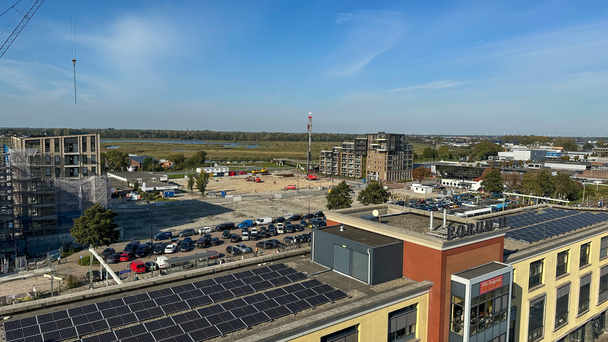 Bouwplaats met meerdere gebouwen in aanbouw, omgeven door parkeerterreinen en een weids landschap met water en bossen in de achtergrond onder een heldere blauwe lucht.