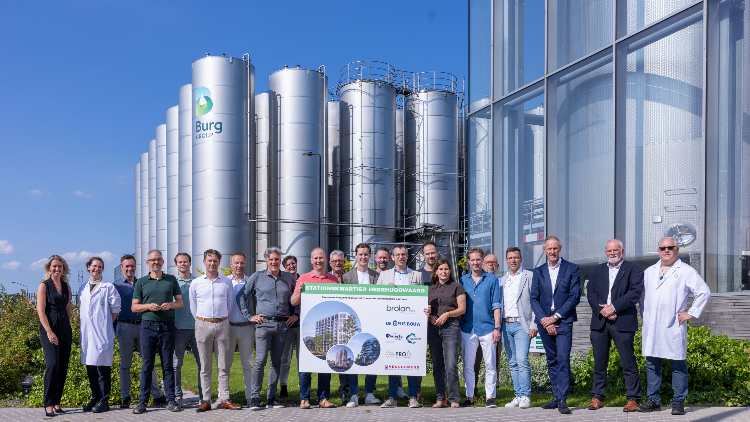 Groep mensen poseert voor een reeks zilverkleurige opslagtanks en een glazen gebouw met het logo van Burg Group. Ze houden een bord vast met de tekst 'Stationskwartier Heerhugowaard' en de namen van verschillende partners. De lucht is helder en blauw.