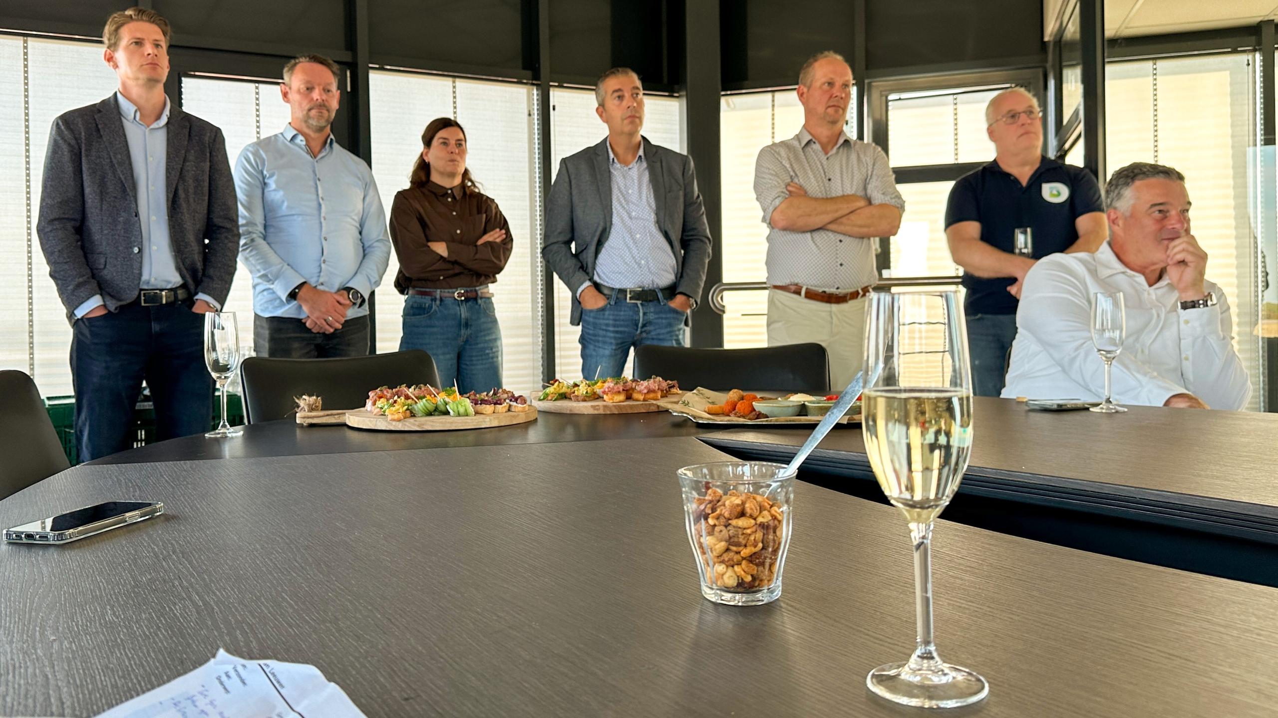 Een groep mensen luistert aandachtig tijdens een bijeenkomst in een moderne ruimte, met hapjes op tafel en een glas champagne op de voorgrond.