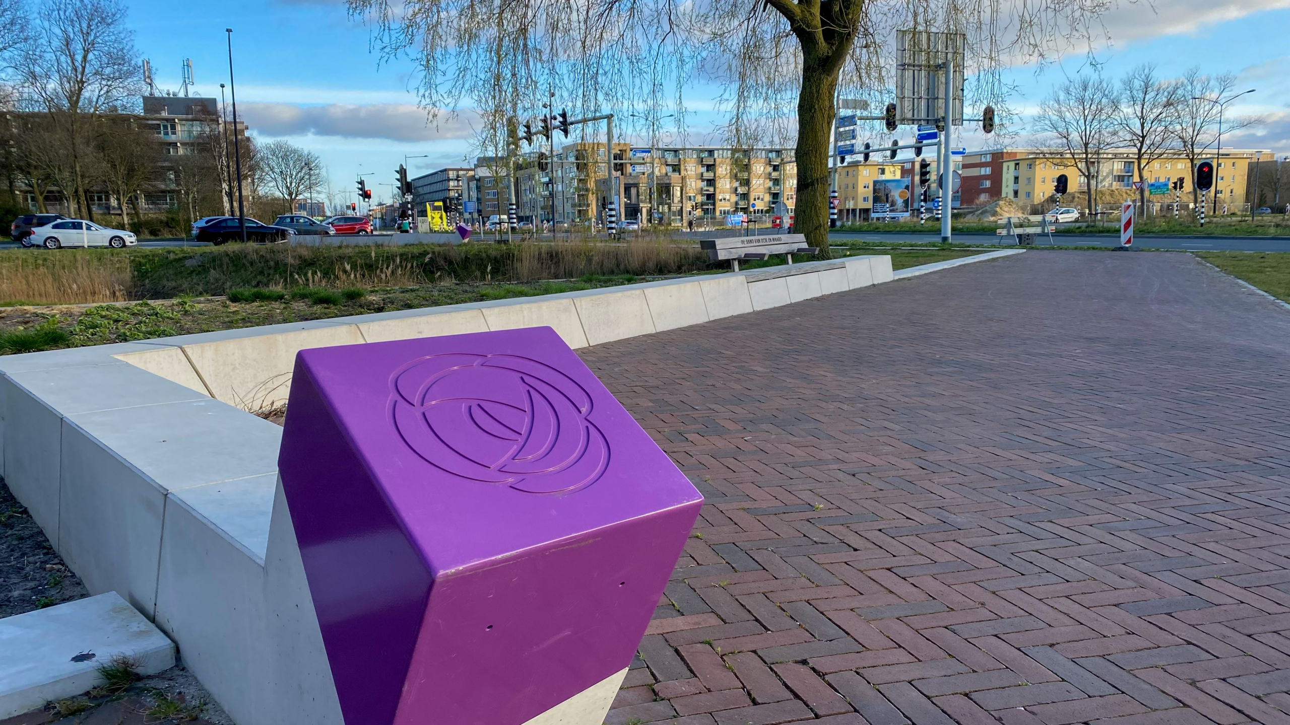 Paarse kubus met een abstract patroon op een betegeld plein, omgeven door groen en een boom, met op de achtergrond auto's, gebouwen en verkeerslichten in een stedelijke omgeving.