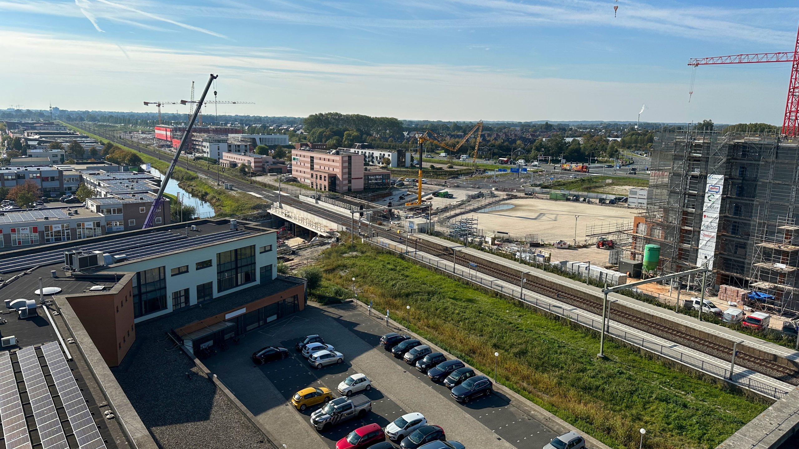 Luchtfoto van een bouwterrein naast een spoorlijn, met meerdere gebouwen en hijskranen in de achtergrond. Er is een parkeerplaats met auto's op de voorgrond, en de omgeving is groen met een heldere lucht.