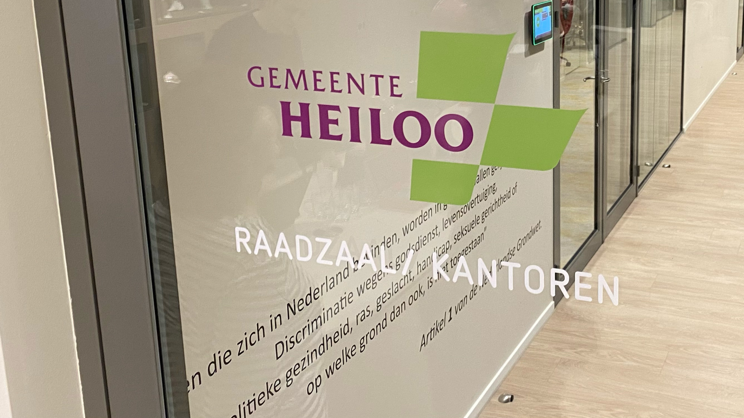 Glazen deur met het logo en de tekst van de gemeente Heiloo, en de woorden 'Raadzaal KANTOREN', met een fragment van artikel 1 van de Nederlandse grondwet over discriminatie daaronder.