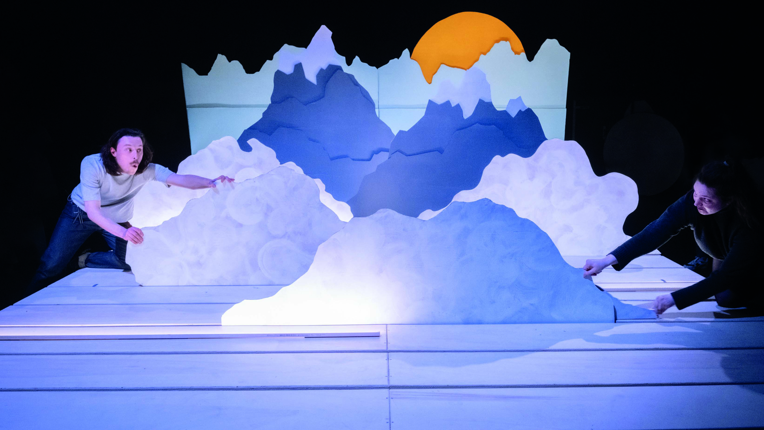 Twee mensen bewegen grote papieren bergen in een theatervoorstelling, met een oranje cirkel die een zon voorstelt op de achtergrond.