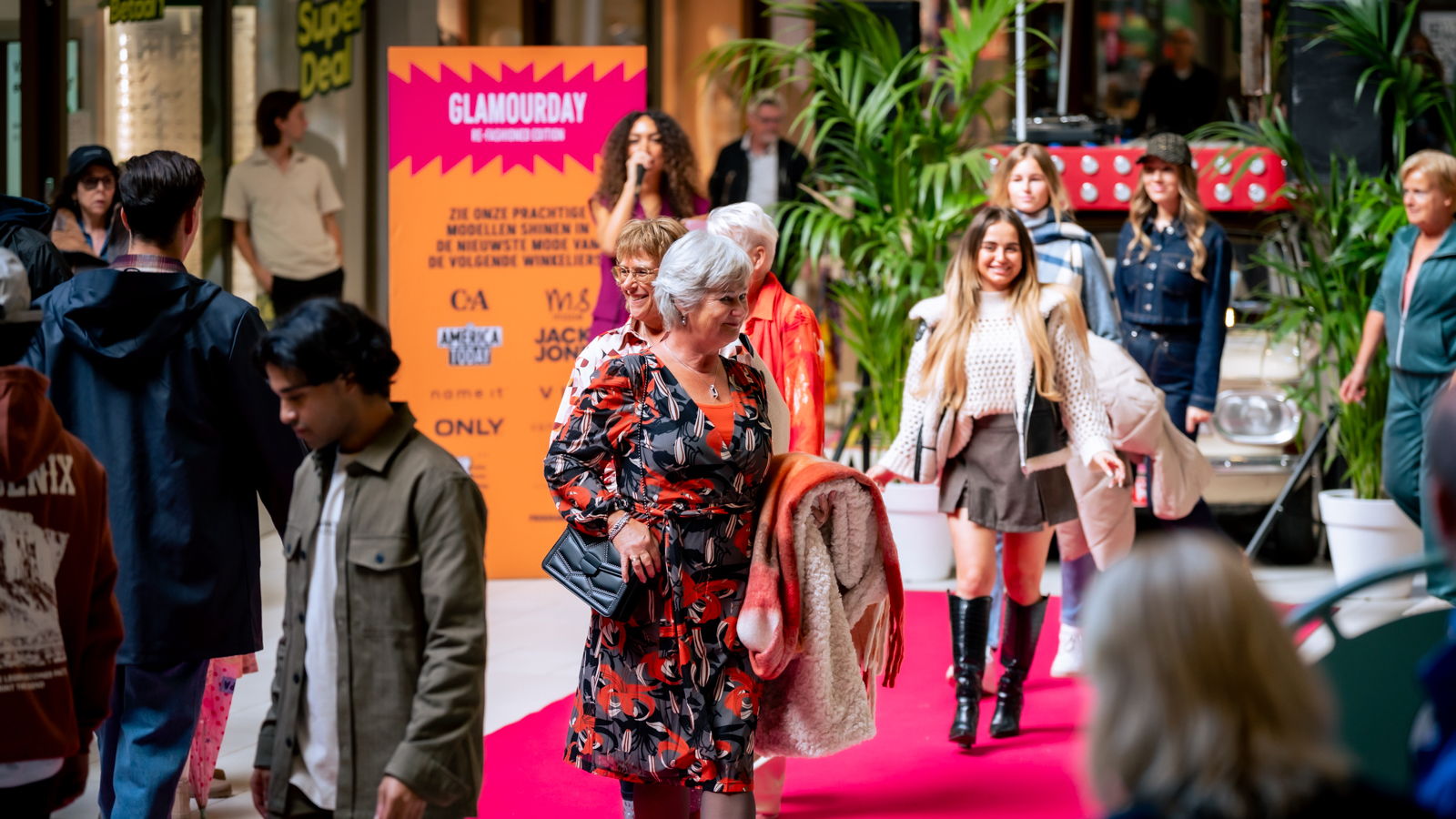 Mensen lopen op een rode loper tijdens een mode-evenement, met een fel oranje bord op de achtergrond met de tekst 'Glamour Day'.