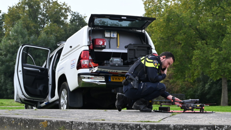 Politieagent knielt naast een groot gestationeerd apparaat op een stenen platform, met een geopende politiewagen op de achtergrond in een groene buitenomgeving.