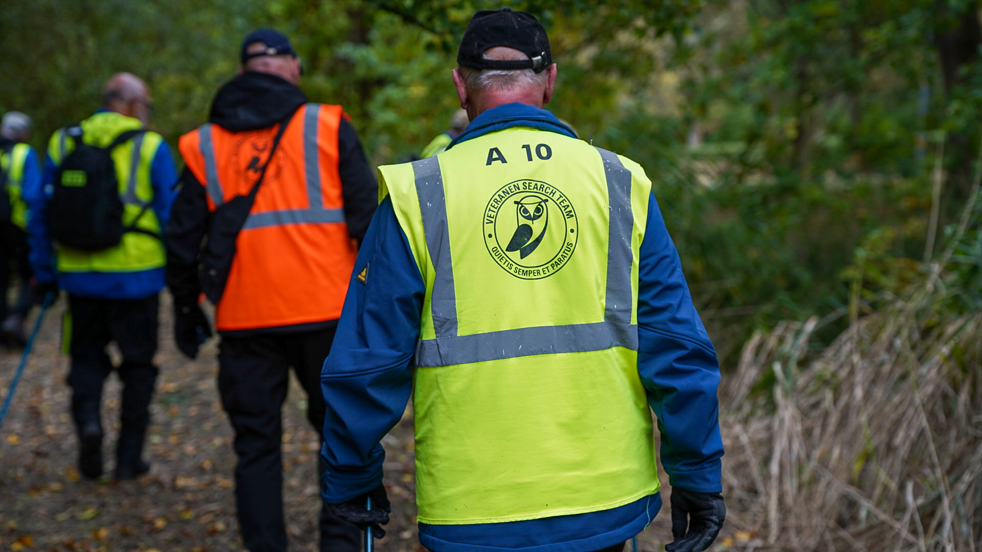 Mensen gekleed in felgekleurde vesten, met de tekst 'Veteranen Search Team', wandelen door een bosrijke omgeving.
