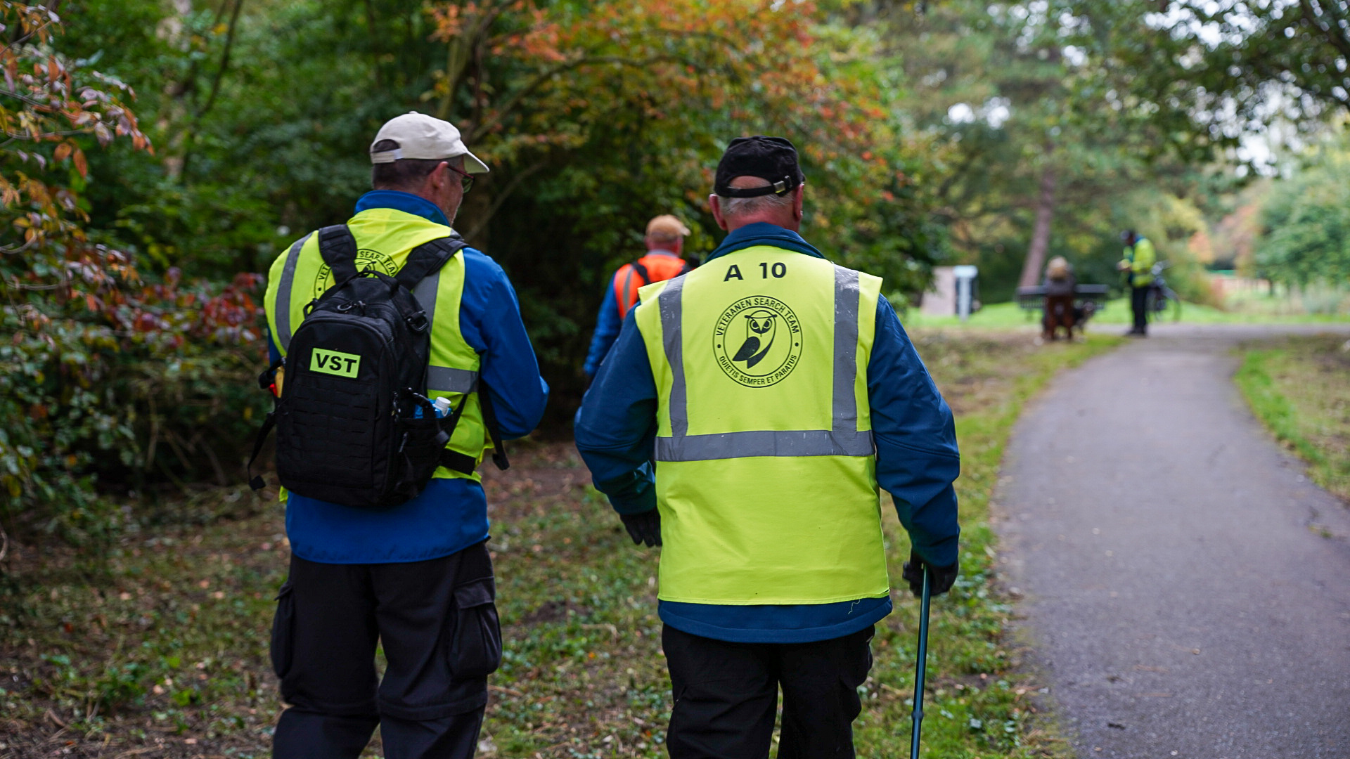 Twee mensen in gele veiligheidshesjes met rugzakken lopen op een pad in een groen park, met andere mensen in de verte.