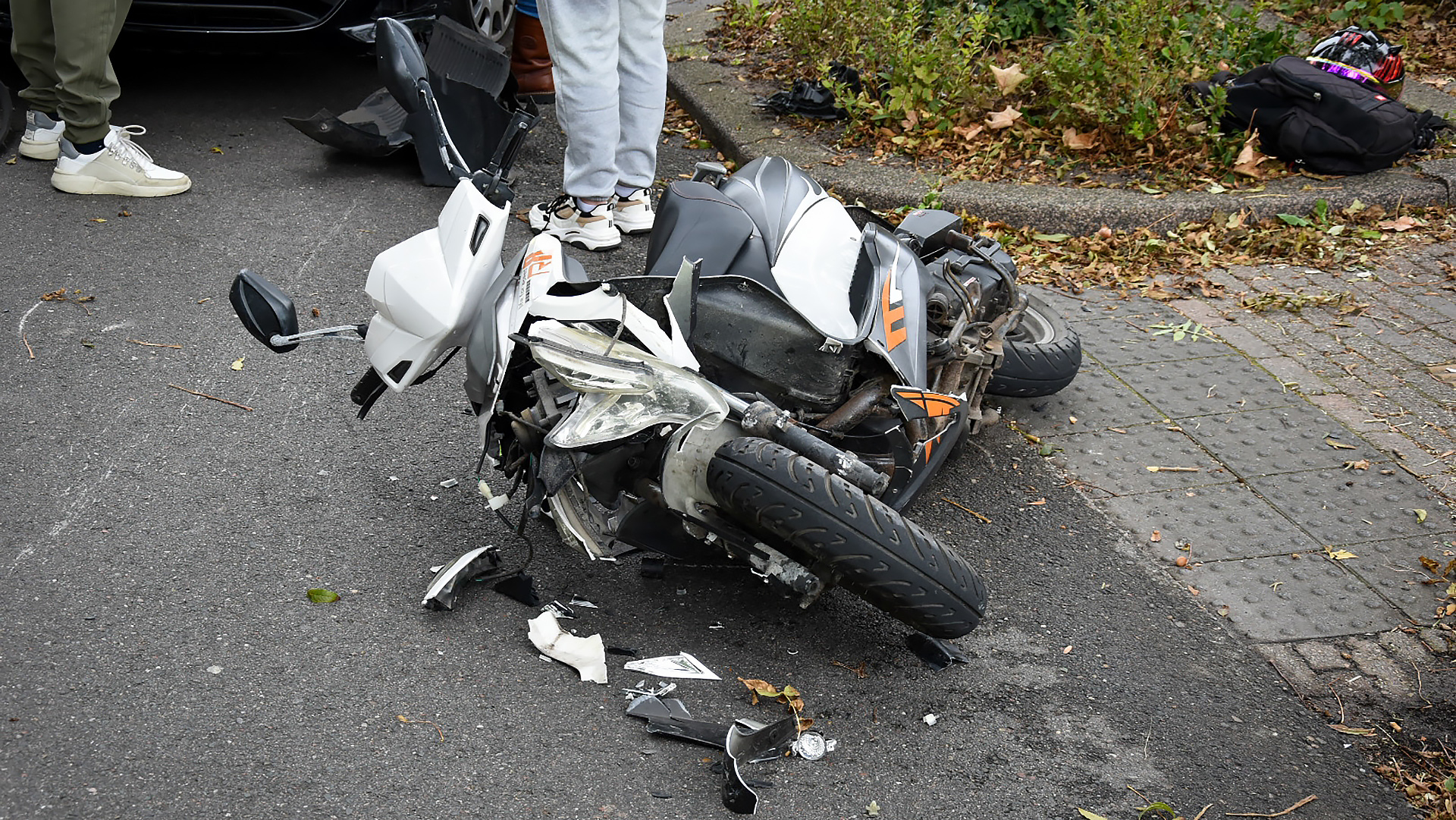Een beschadigde scooter ligt op de grond naast een trottoir, met stukken van de scooter verspreid eromheen. Drie paar benen van mensen zijn zichtbaar, samen met een beschadigde auto op de achtergrond.