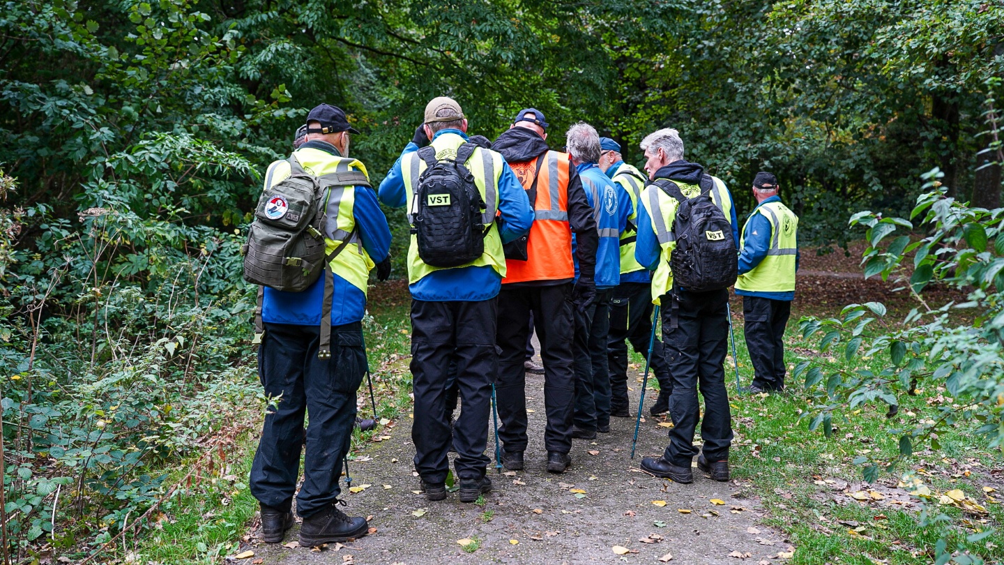 Een groep mensen in felgekleurde hesjes en rugzakken staat samen op een bos pad, omringd door groene bomen en struiken.
