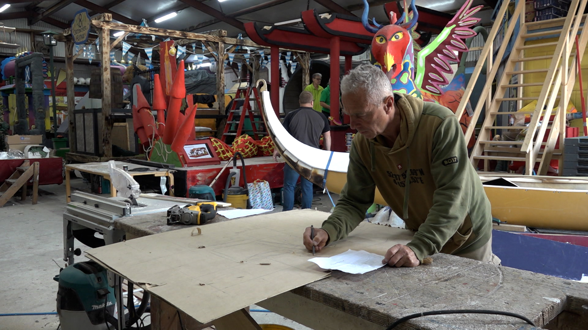 Man bezig met een tekening op een werkbank in een grote werkplaats, omgeven door kleurrijke decorstukken en gereedschap.