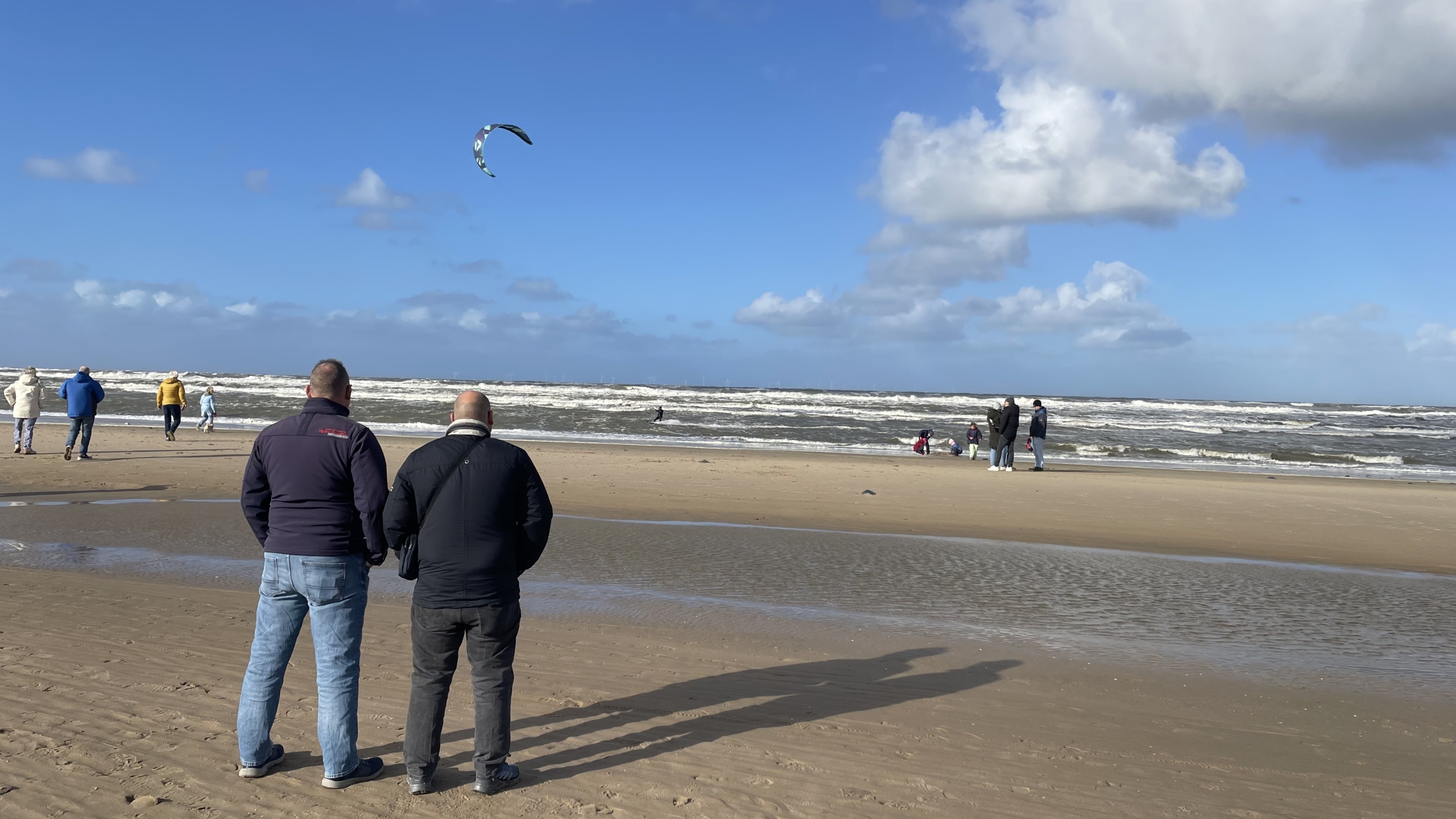Een zonnig strand met een aantal mensen die langs de waterkant lopen en één persoon die aan het kitesurfen is, met zichtbare wolken en een blauwe hemel.