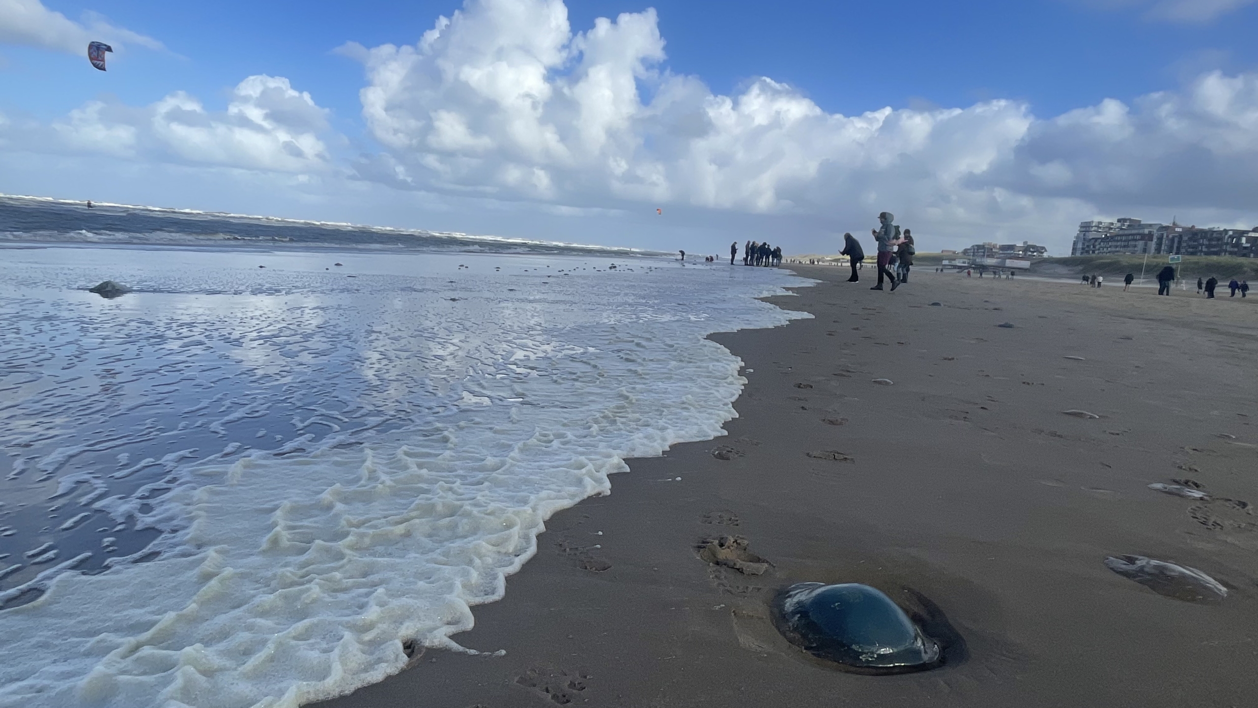 Strand met witte schuimvlokken op het zand en mensen die langs de kustlijn lopen onder een blauwe hemel met wolken.