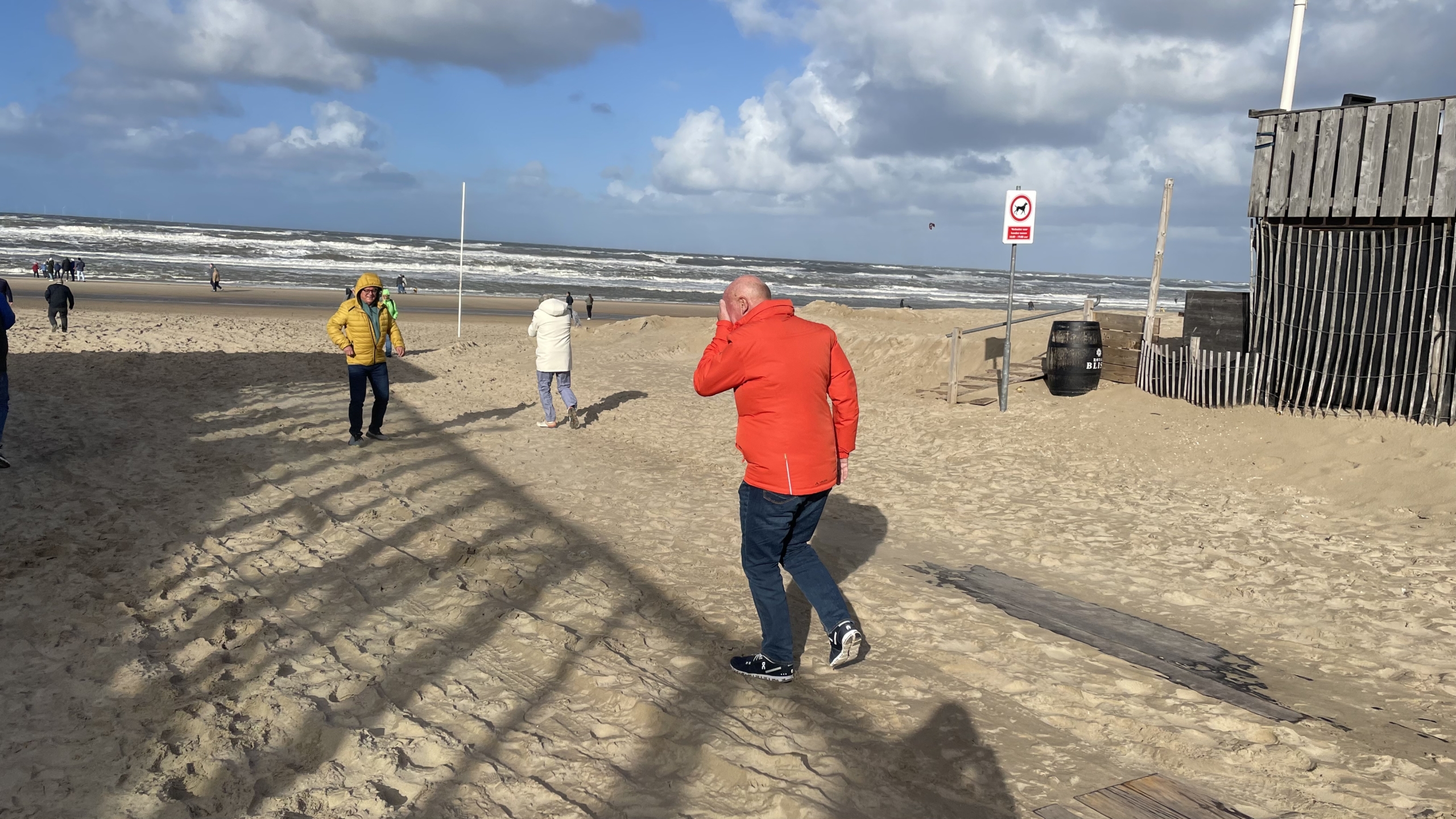 Mensen wandelen op een winderig strand met hoge golven en bewolkte lucht terwijl ze warme jassen dragen; een persoon loopt tegen de wind in met een rode jas aan, naast een gebouw en een verbodsbord op het strand.