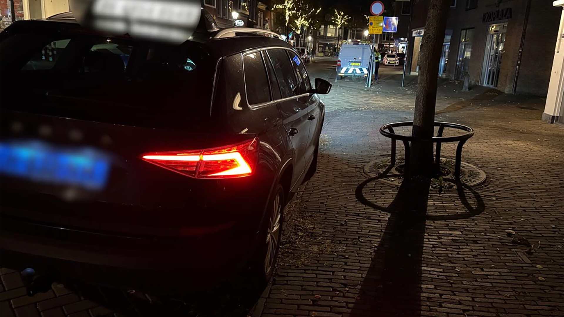 Een zwarte auto geparkeerd op een stoep naast een boom in een verlichte straat 's nachts, met brandende achterlichten en onleesbare kentekenplaten.