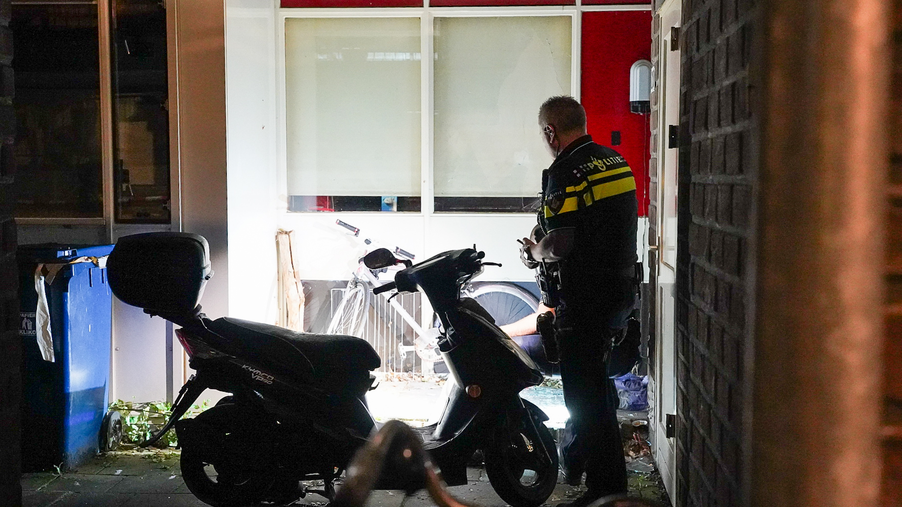 Een politieagent staat bij een scooter in een smalle steeg met een blauwe afvalcontainer aan de linkerkant. De agent lijkt aantekeningen te maken. Op de achtergrond zijn een fiets en een gebouw met een rode en witte gevel zichtbaar.