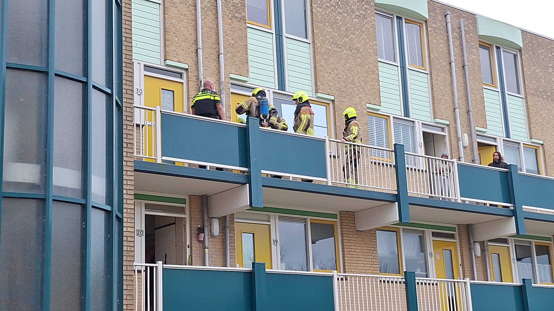 Brandweerlieden en een politieagent staan op een balkon van een appartementencomplex. Ze lijken bezig met een noodsituatie of inspectie. Er zijn ook andere mensen op het balkon en aan het raam te zien.
