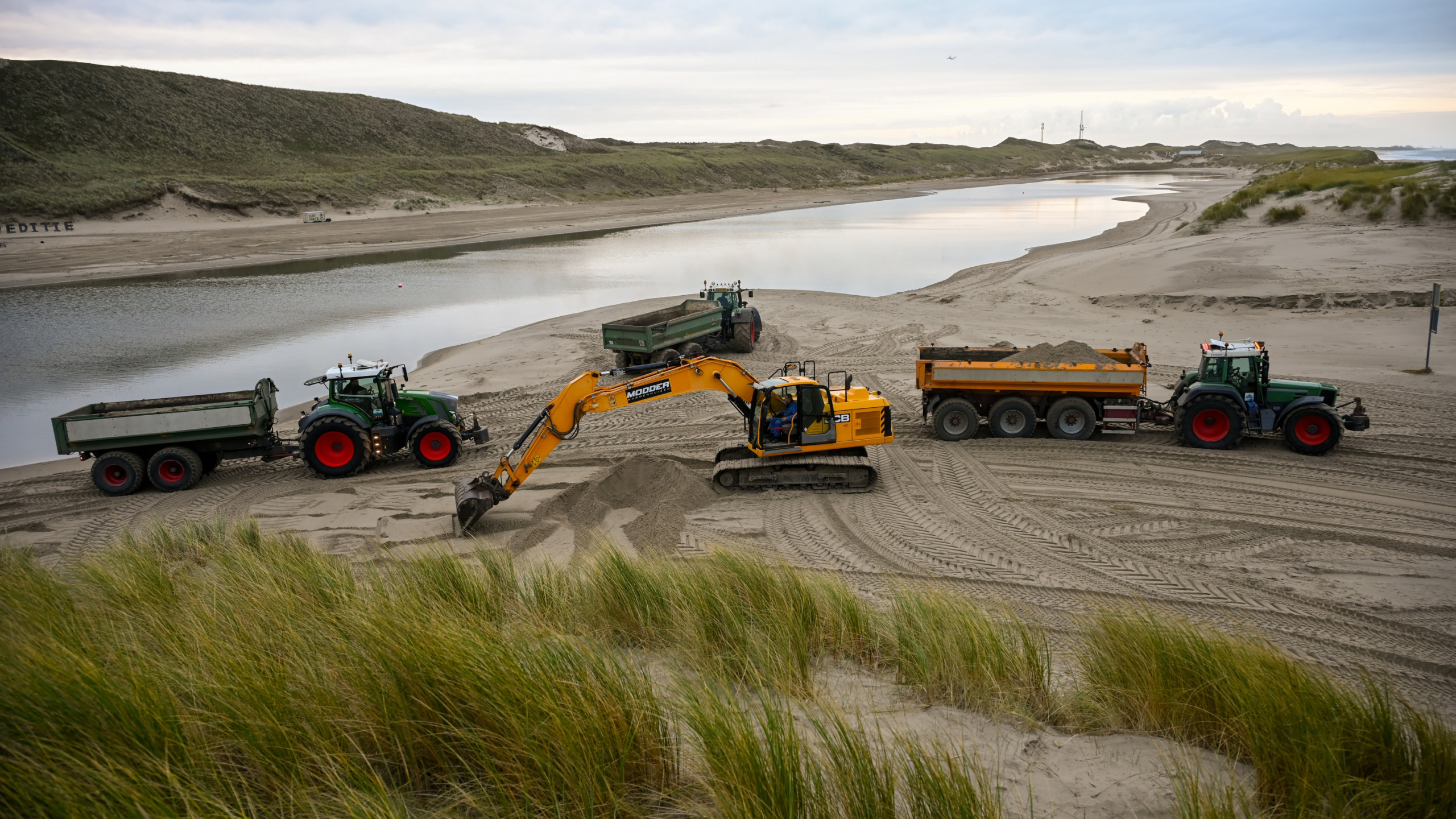 Graafmachines en tractoren voeren zandverplaatsingen uit op een strand, met een duinlandschap en water op de achtergrond.
