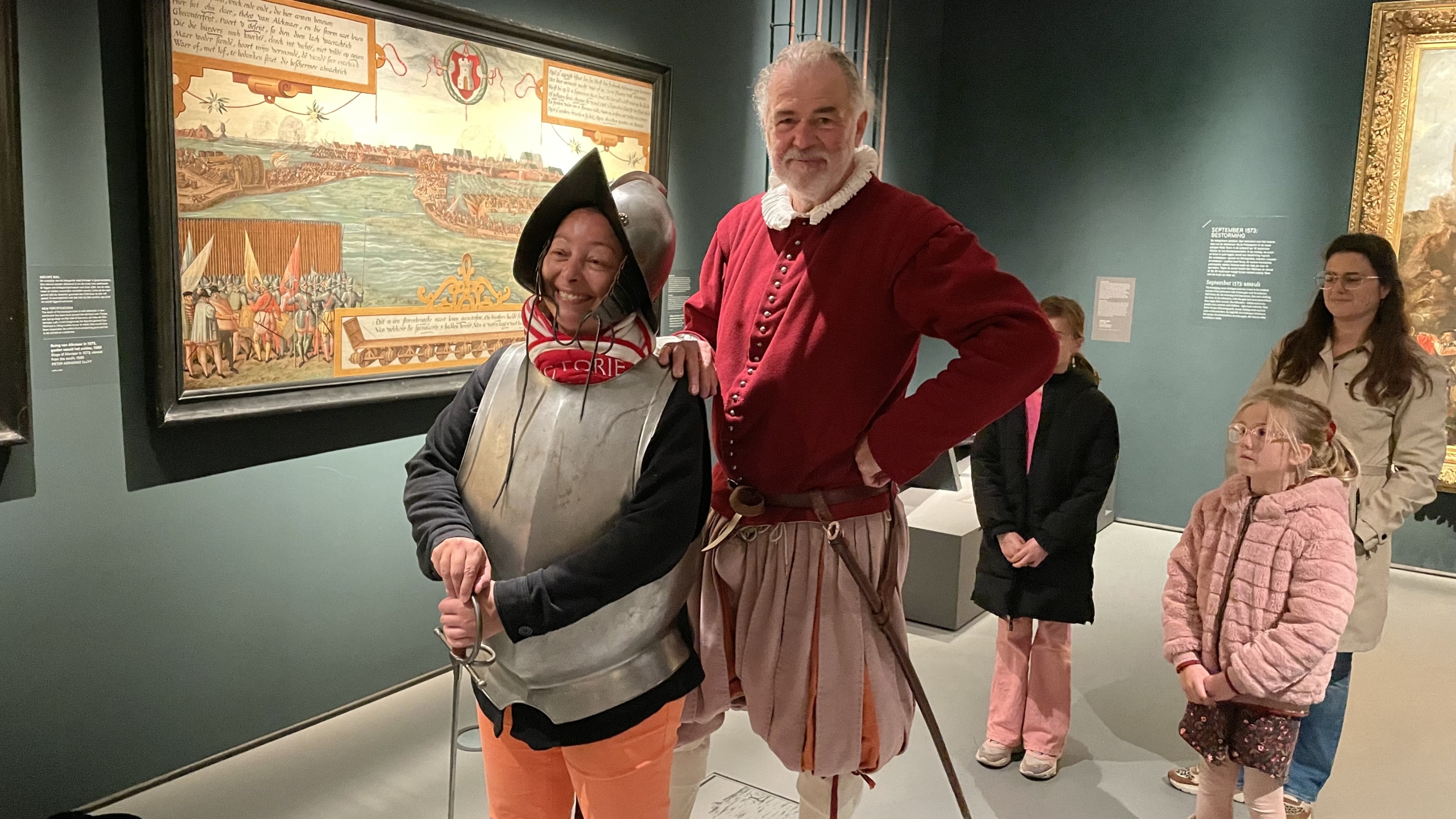 Een persoon in een harnas met helm, en een andere persoon in historische kleding poseren voor een schilderij in een museum, met enkele toeschouwers op de achtergrond.