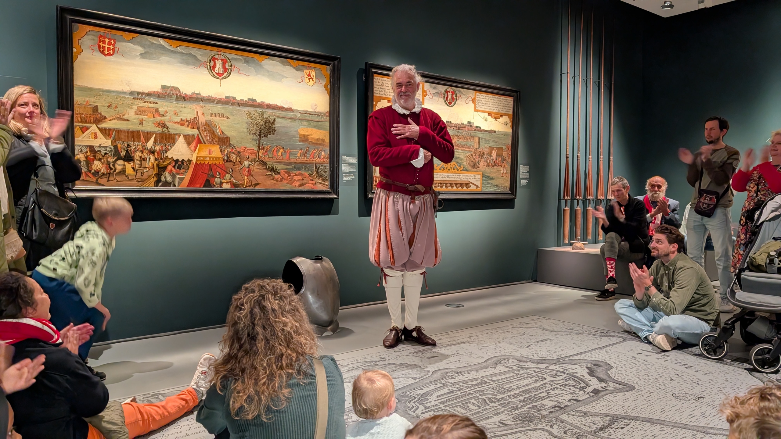 Een man in een historisch kostuum staat voor een publiek in een museum, omgeven door schilderijen aan de muur, terwijl het publiek applaudisseert.