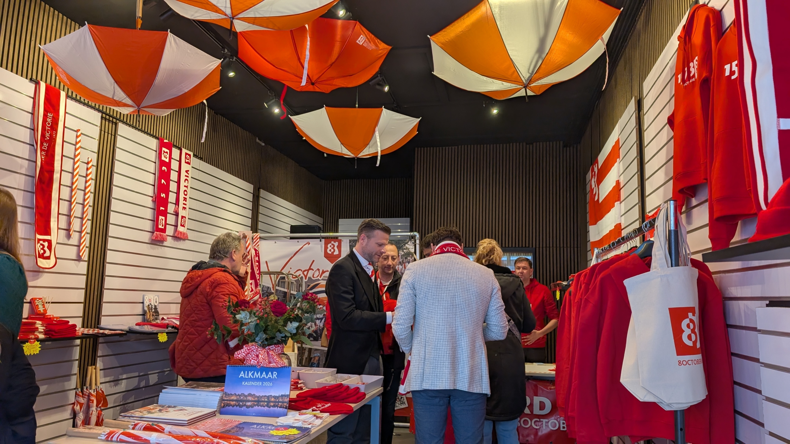Mensen in een winkel met rood-witte merchandise, waaronder sjaals en truien, met rode en witte parasols aan het plafond.