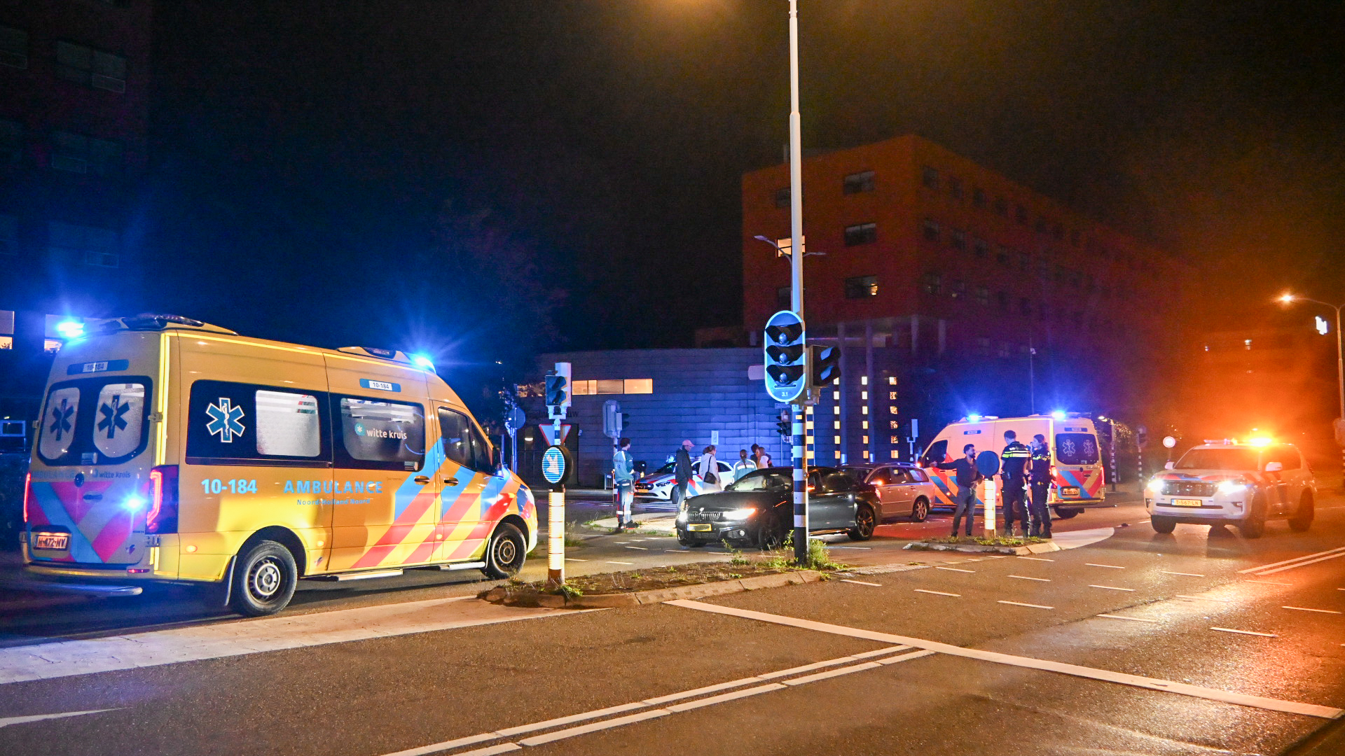 Politievoertuigen, ambulances en mensen bij een verkeersongeluk op een kruispunt 's nachts met blauwe en rode lichten die de omgeving verlichten.