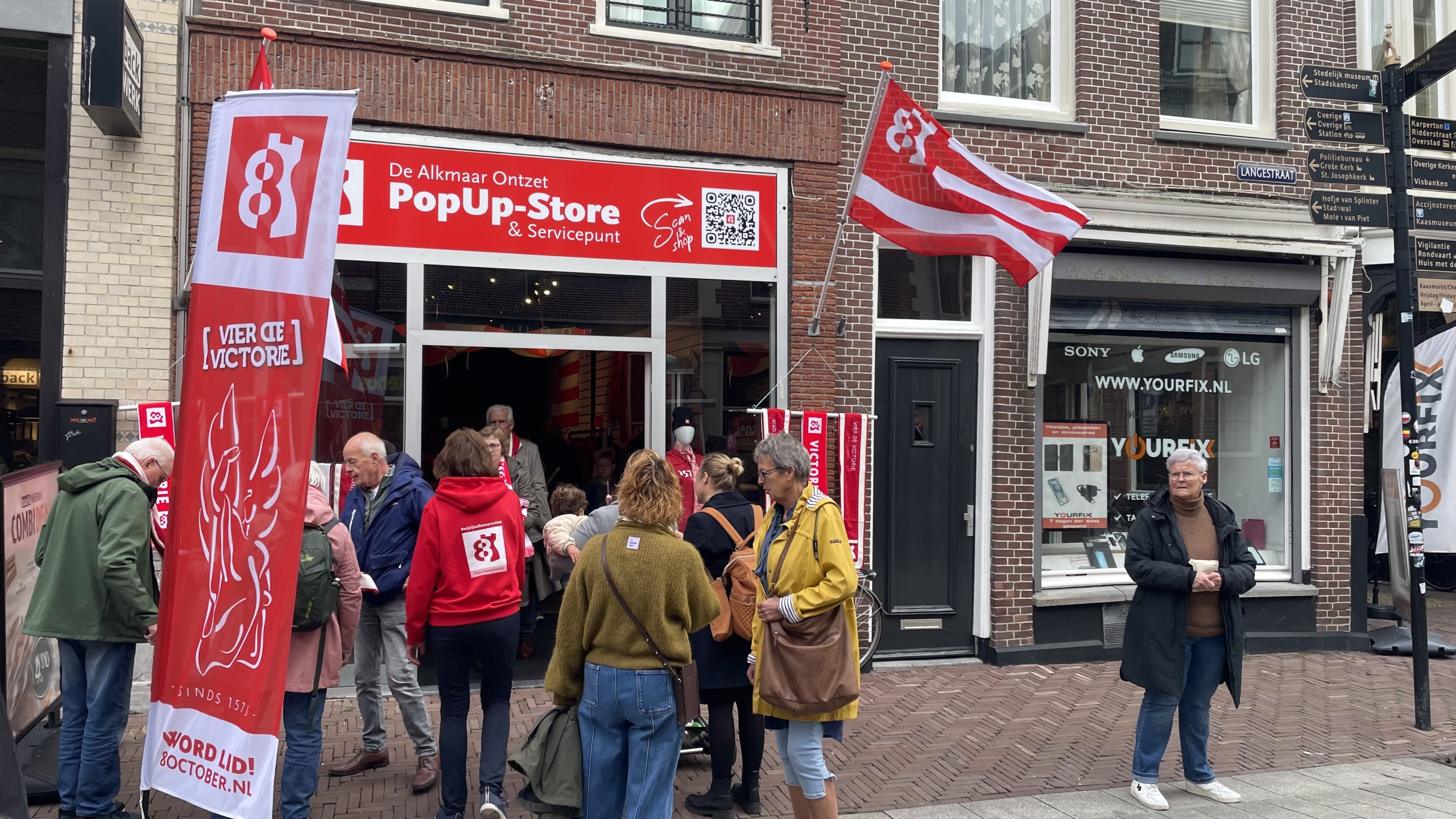 Mensen verzamelen zich voor de Alkmaar Ontzet Pop-Up Store in een straat met rode en witte decoraties en vlaggen.