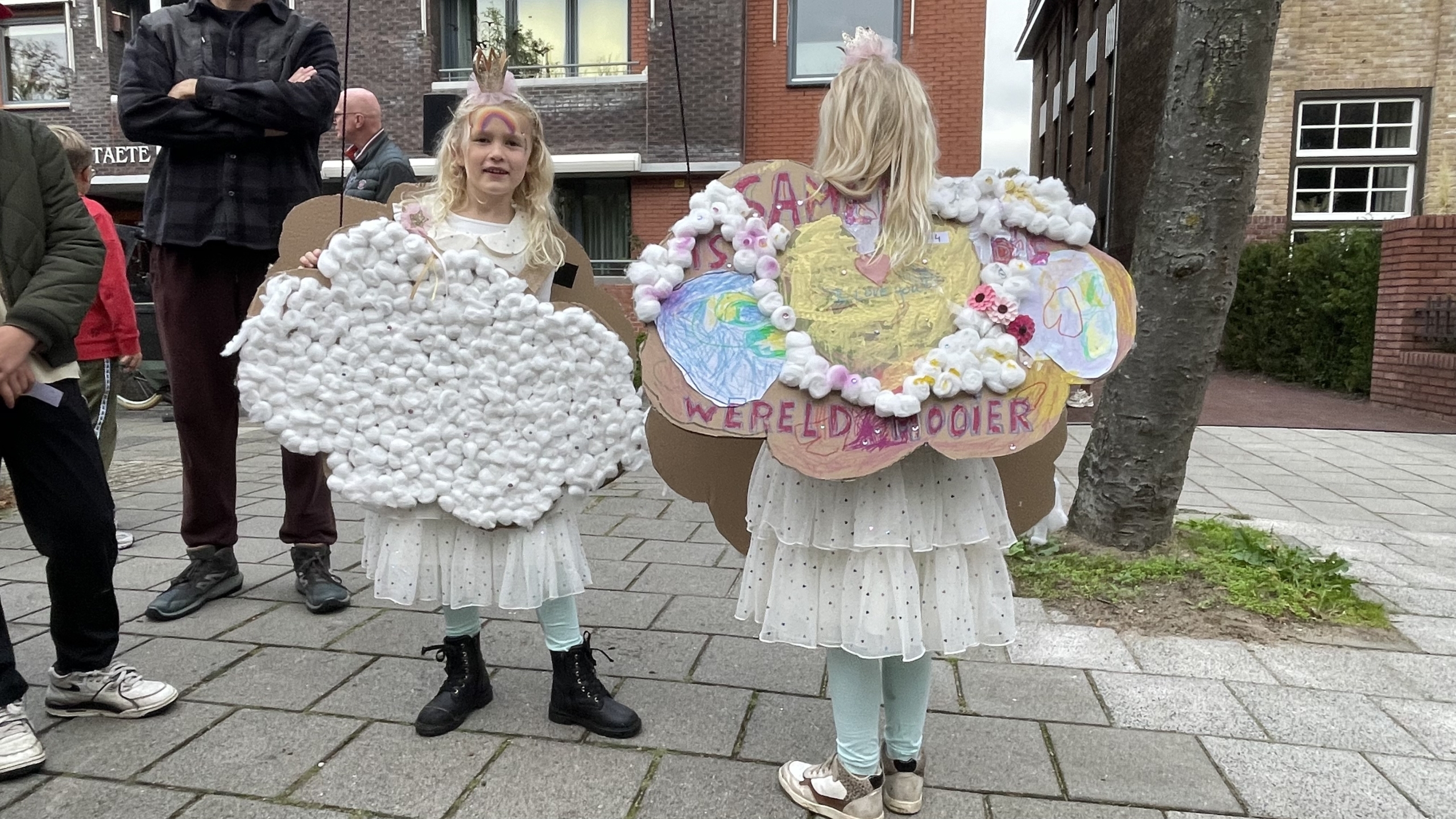 Twee jonge meisjes in witte outfits staan op een trottoir, elk met een zelfgemaakt kartonnen kunstwerk versierd met wattenbolletjes en kleurrijke tekeningen. Andere mensen staan op de achtergrond, waaronder een man in donkere kleding met gekruiste armen. Eén meisje draagt een hoofddeksel met een kroon erop.