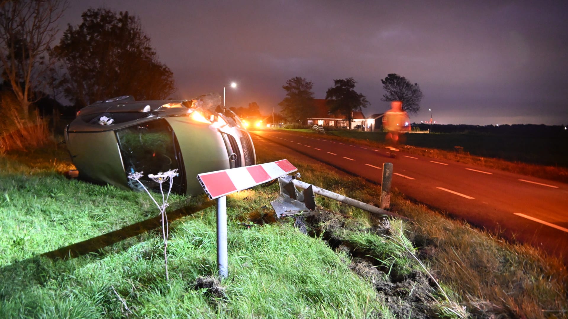 Een omgevallen auto ligt op zijn kant naast een landelijke weg in het gras, met een beschadigd verkeersbord op de voorgrond en een persoon die in de verte loopt bij schemering.