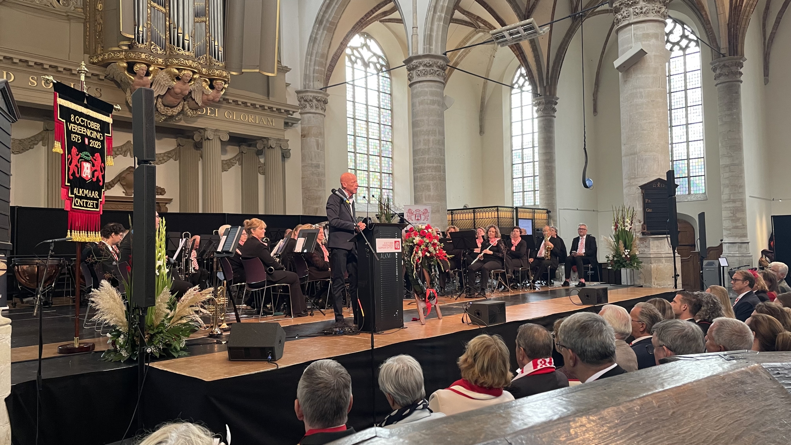 Een spreker staat op een podium in een historische kerk, met een orkest zittend op de achtergrond. Er hangt een vlag met de tekst "8 Oktober Vereniging 1573-2023 Alkmaar Ontzet". Er zijn toeschouwers die aandachtig luisteren.