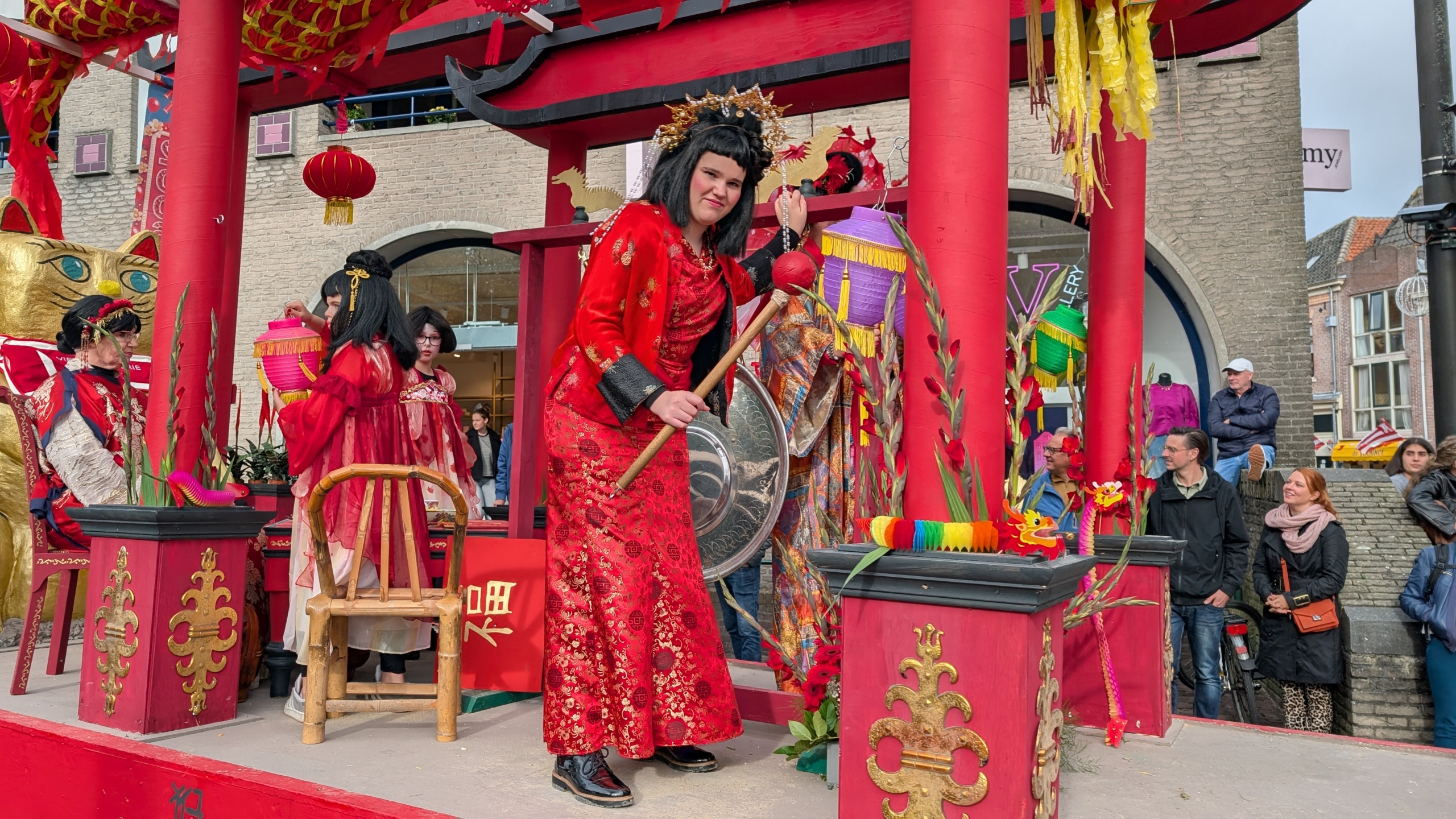 Mensen in traditionele kleding poseren op een sierlijke, rode wagen versierd met Chinese draken en een grote gouden kat tijdens een parade, terwijl omstanders toekijken.