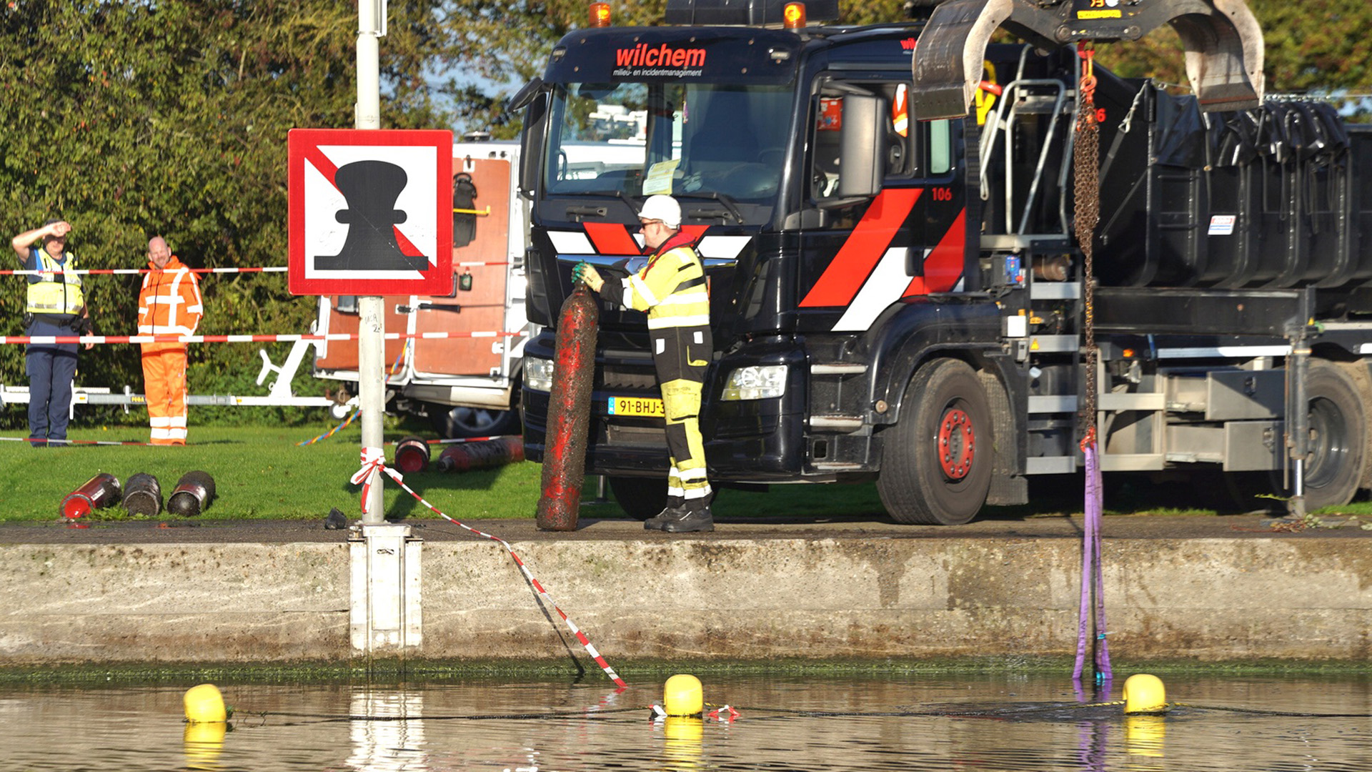 Een man in veiligheidskleding staat naast een vrachtwagen met de tekst "wilchem" en houdt een groot, cilindervormig object vast. Er zijn meer mensen in oranje veiligheidskleding te zien op de achtergrond. Een bord met een afbeelding van een meerpaal die doorstreept is, staat op de voorgrond. Er is water met gele boeien op de voorgrond te zien.