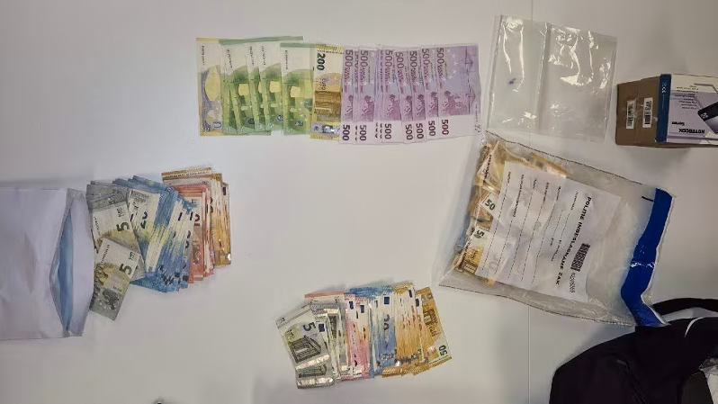 Verschillende stapels eurobiljetten in diverse coupures liggen verspreid op een witte tafel, naast een geopende envelop en een afgesloten plastic zak met documenten.