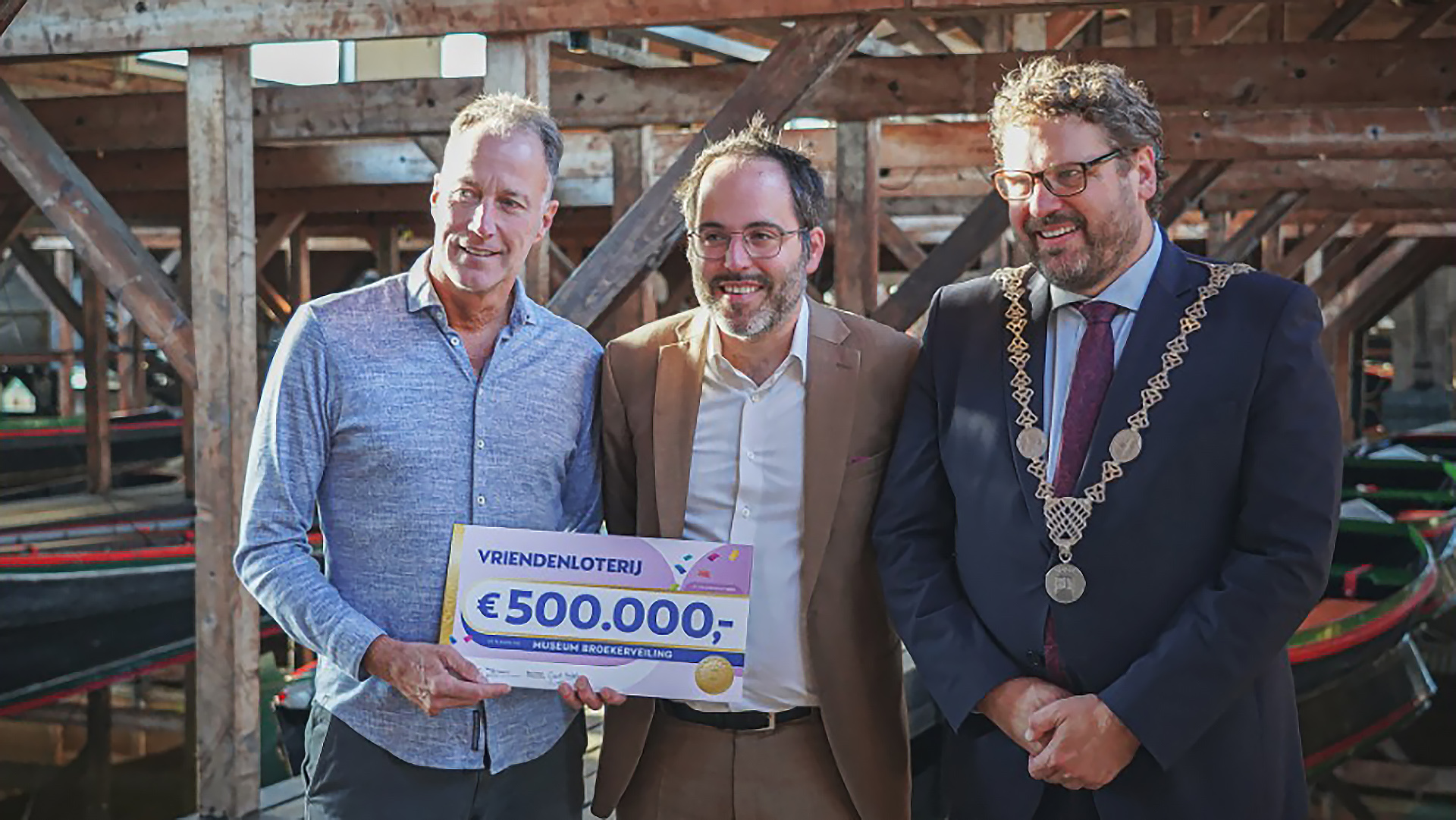 Drie mannen poseren met een grote cheque van 500.000 euro van de Vriendenloterij in een ruimte met houten balken.