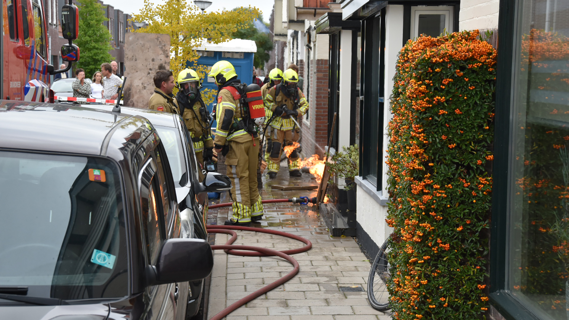 Brandweerlieden in volledige beschermende uitrusting bestrijden een vuur aan de kant van een straat, naast een rij huizen met een vrolijke oranje bloeiende haag.