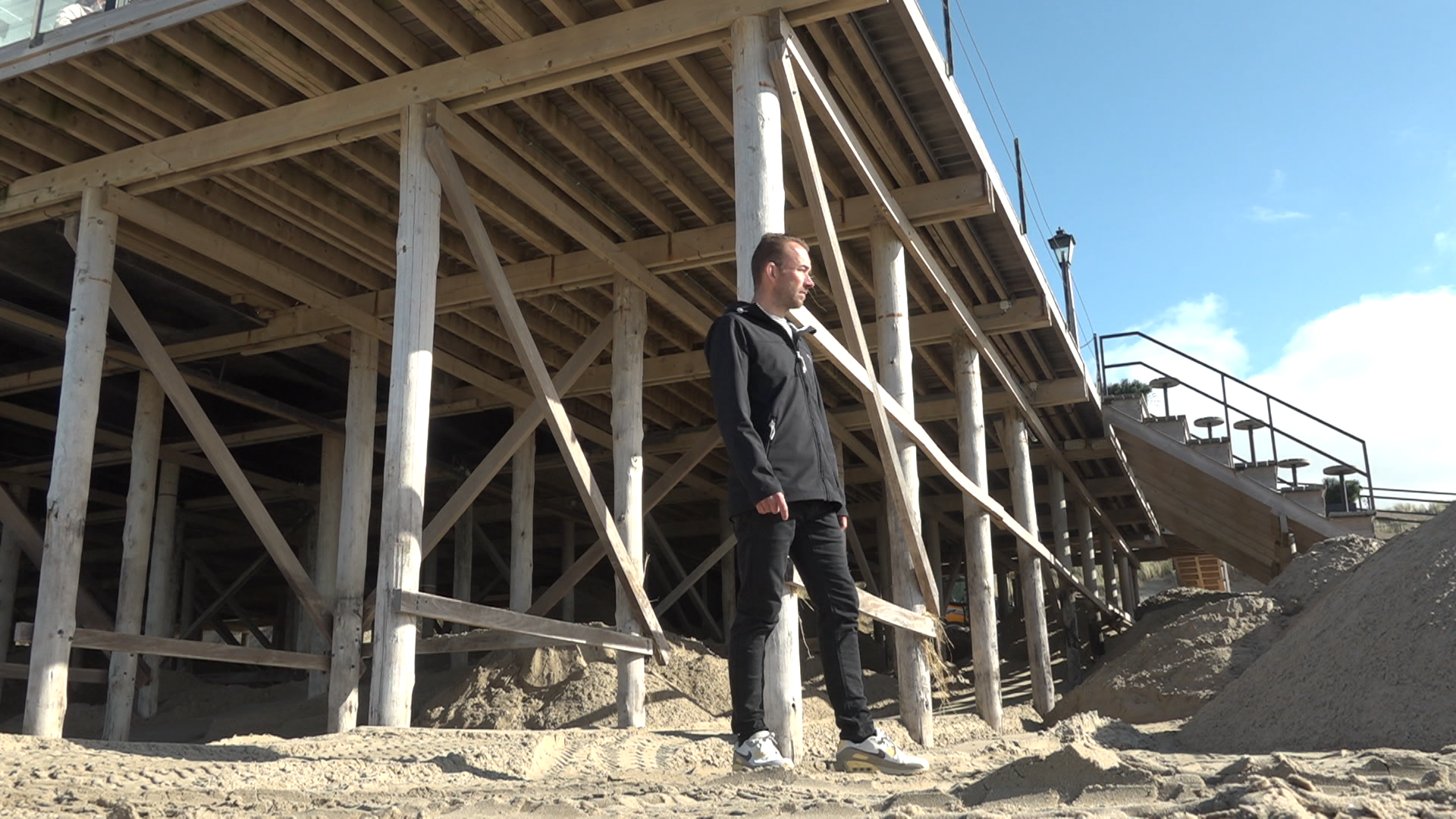 Een man in een zwarte jas en schoenen staat op het zand onder een houten constructie met palen en trappen op een strand.