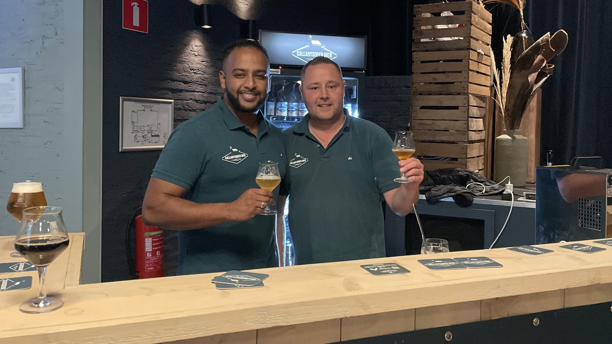 Twee mannen in groene polo's staan achter een houten bar en houden glazen bier vast, met op de achtergrond een koelkast en decoratieve planten.