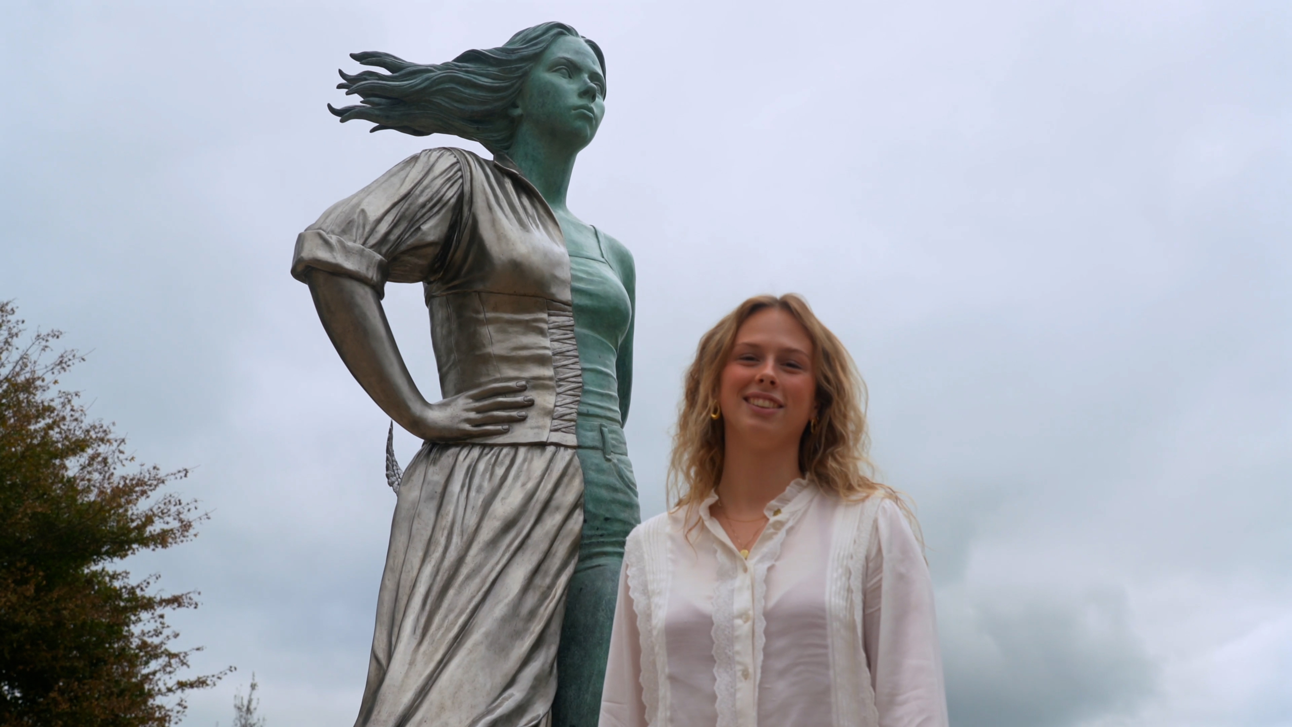 Een jonge vrouw in een witte blouse staat naast een groot standbeeld van een vrouw met wapperend haar in de wind, onder een bewolkte hemel.