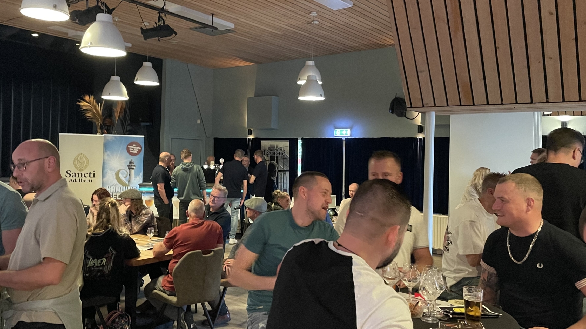 Groep mensen in een gezellige binnenruimte met tafels en stoelen, pratend en drinken, onder een plafond met houten panelen en ronde hanglampen.