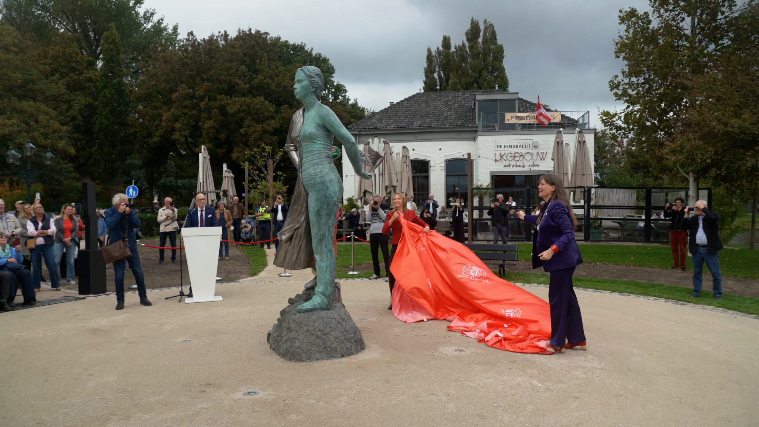 Een bronzen standbeeld wordt onthuld door twee vrouwen die een rood kleed verwijderen, terwijl een groep mensen toekijkt.