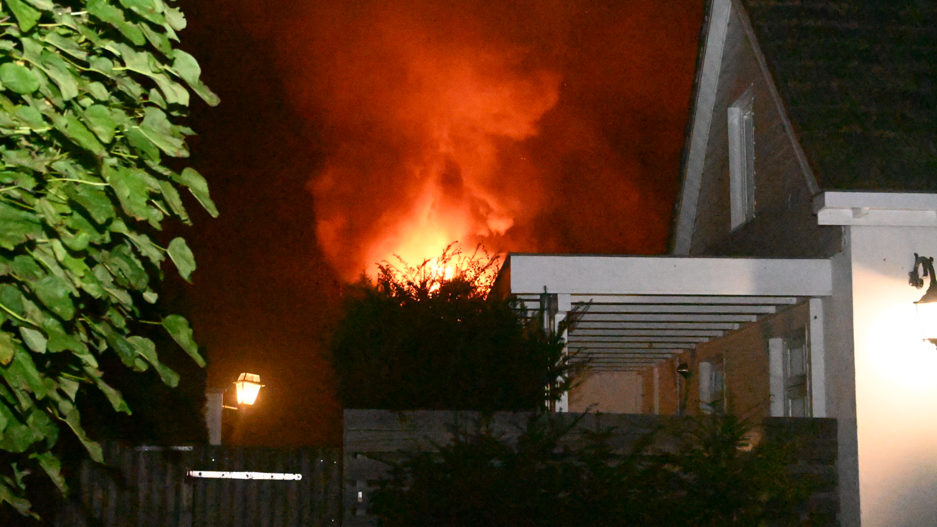 Nachtelijke scène van een brand waarbij vlammen uit het dak komen van een huis, met rook die de donkere lucht vult, en een deel van het huis en de tuin zichtbaar.
